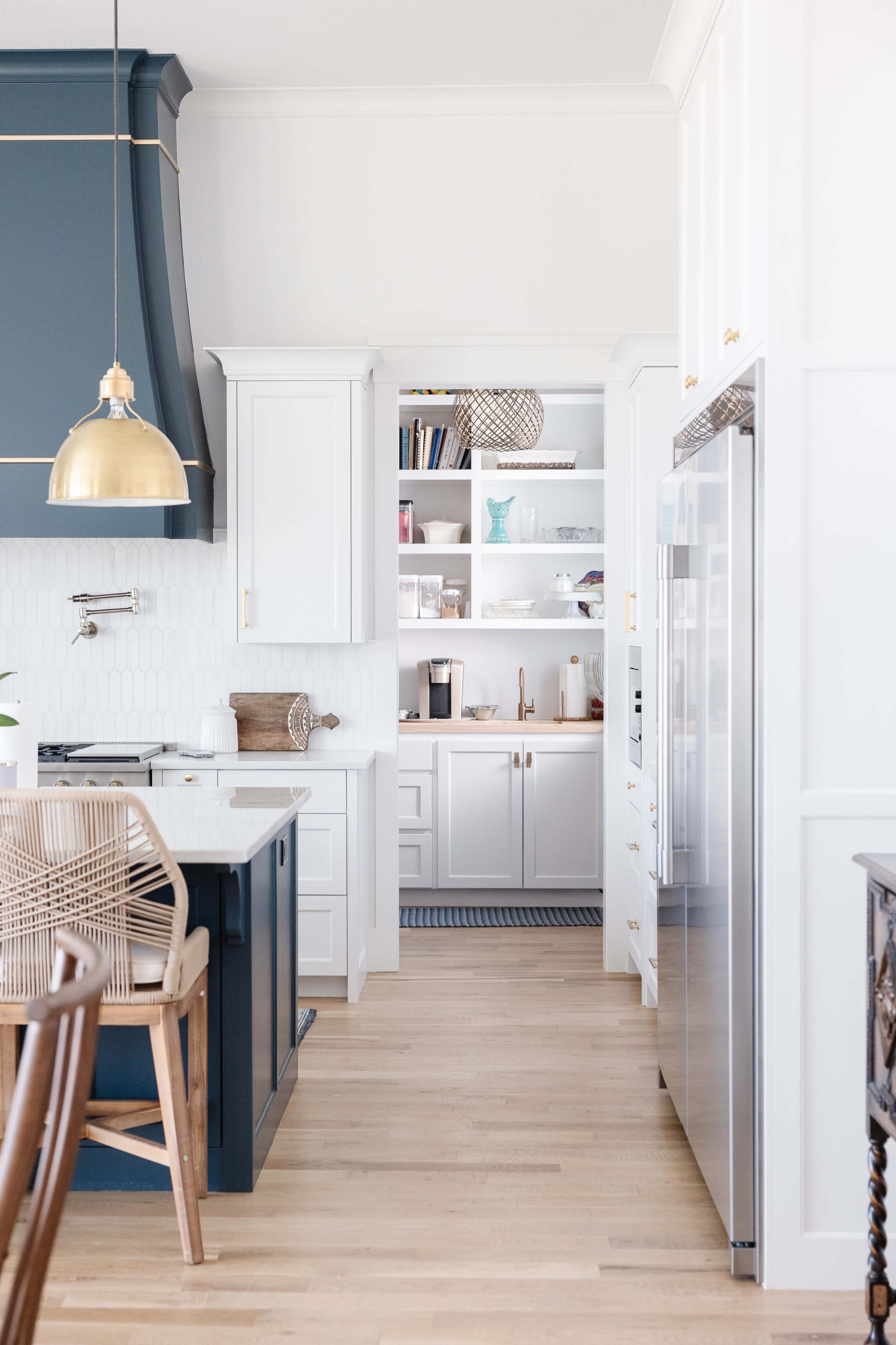 The image shows a modern kitchen featuring a blue and white color scheme, with a large island, a gold pendant light, and an open pantry visible in the background.