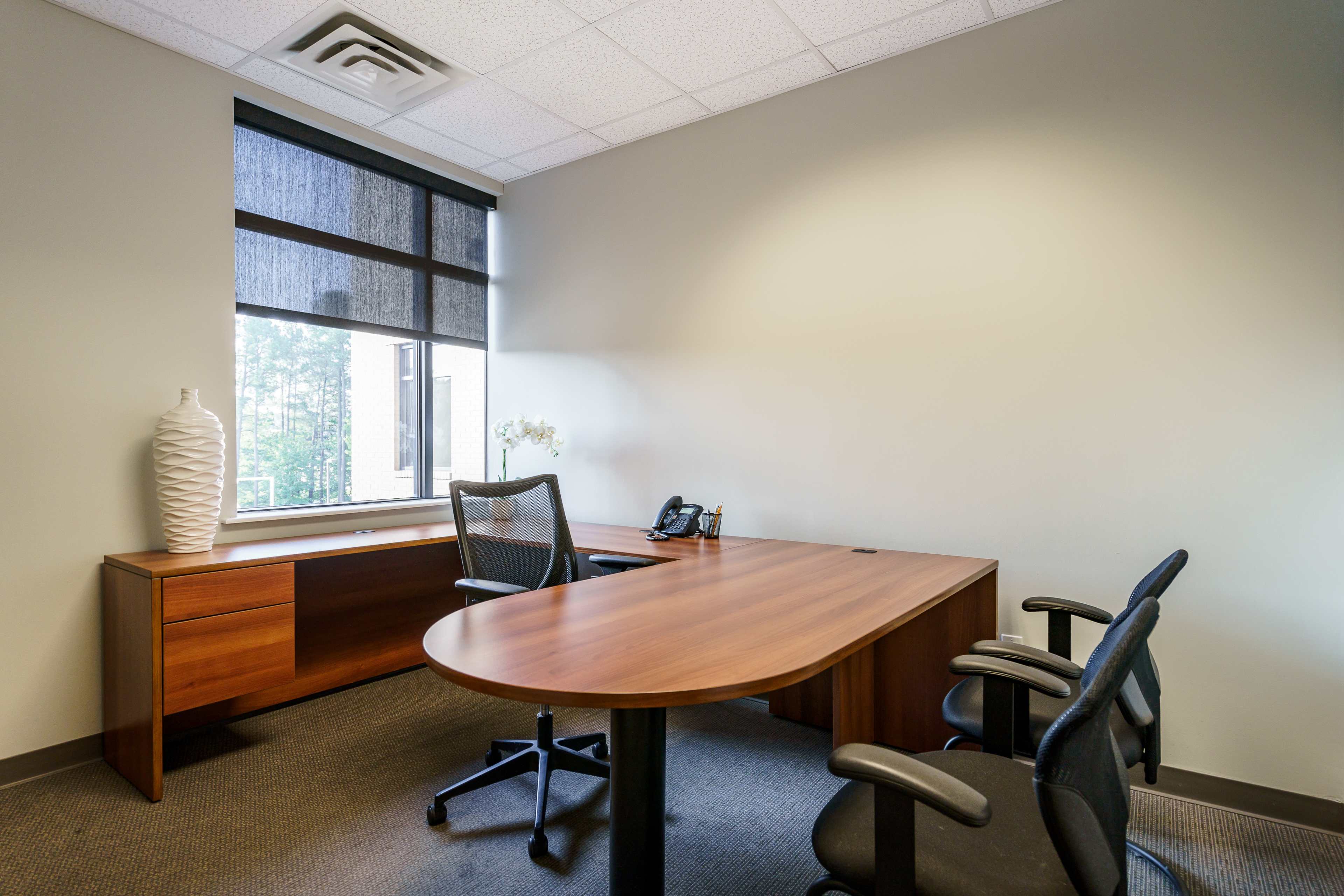 An office space features a wooden desk and chair set, a black telephone, and a window with a roller shade.