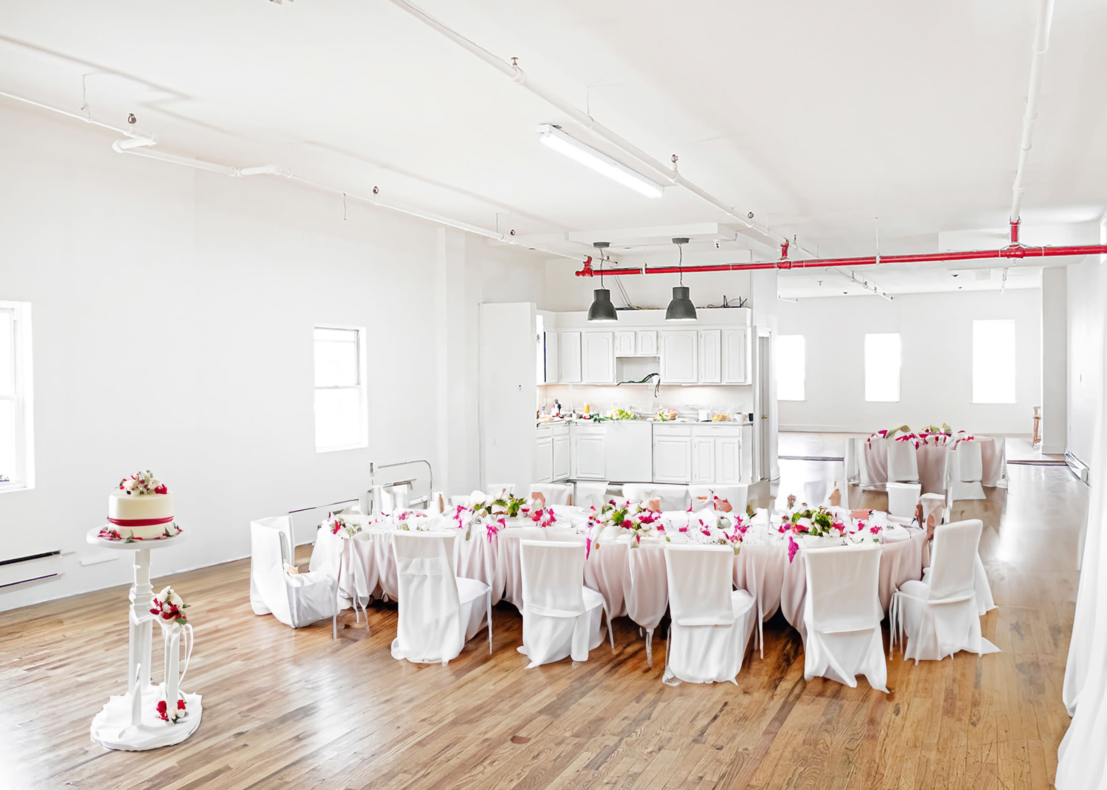 A large, elegantly set table with white tablecloths and floral centerpieces occupies a bright, open space with wooden floors and white walls.