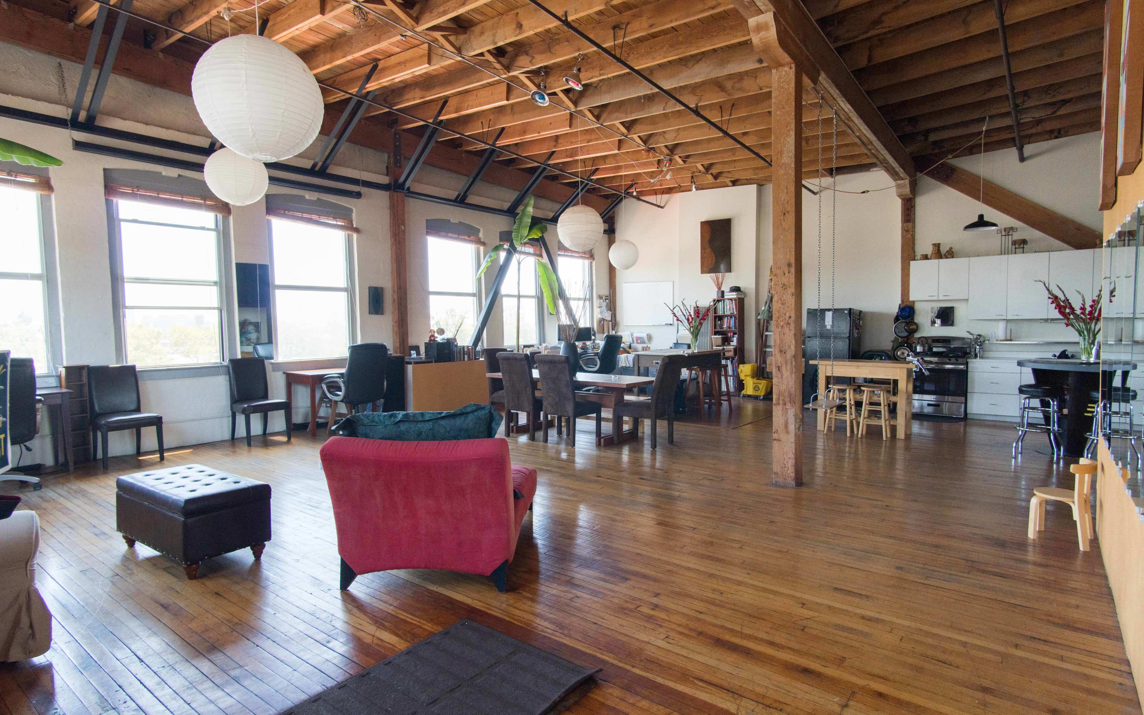 An open-plan loft features wooden floors, high ceilings with exposed beams, and a mix of seating areas along with a dining and kitchen space.