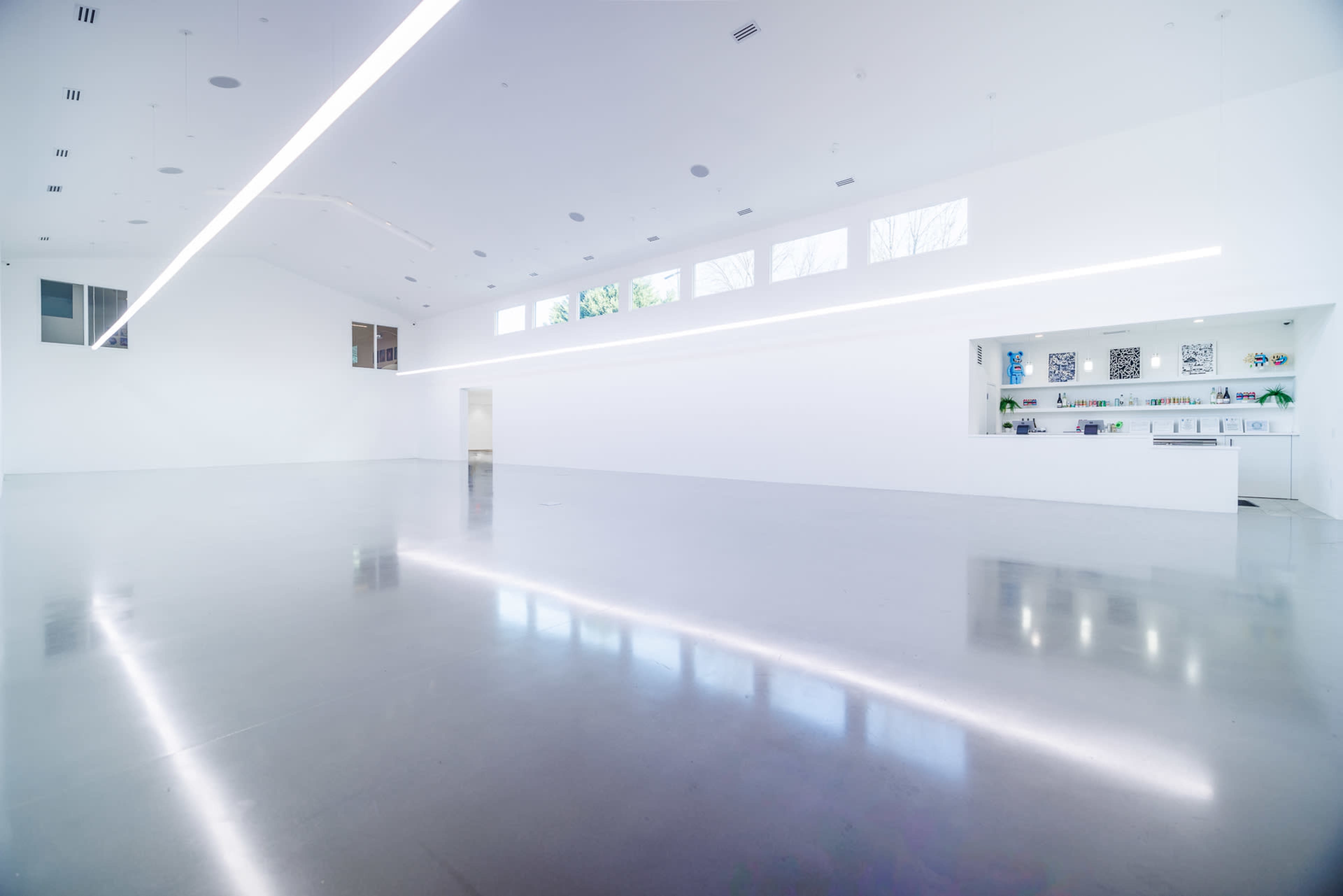 The image shows a spacious, minimalist interior with white walls, polished concrete flooring, and a long counter with artwork displayed on it.