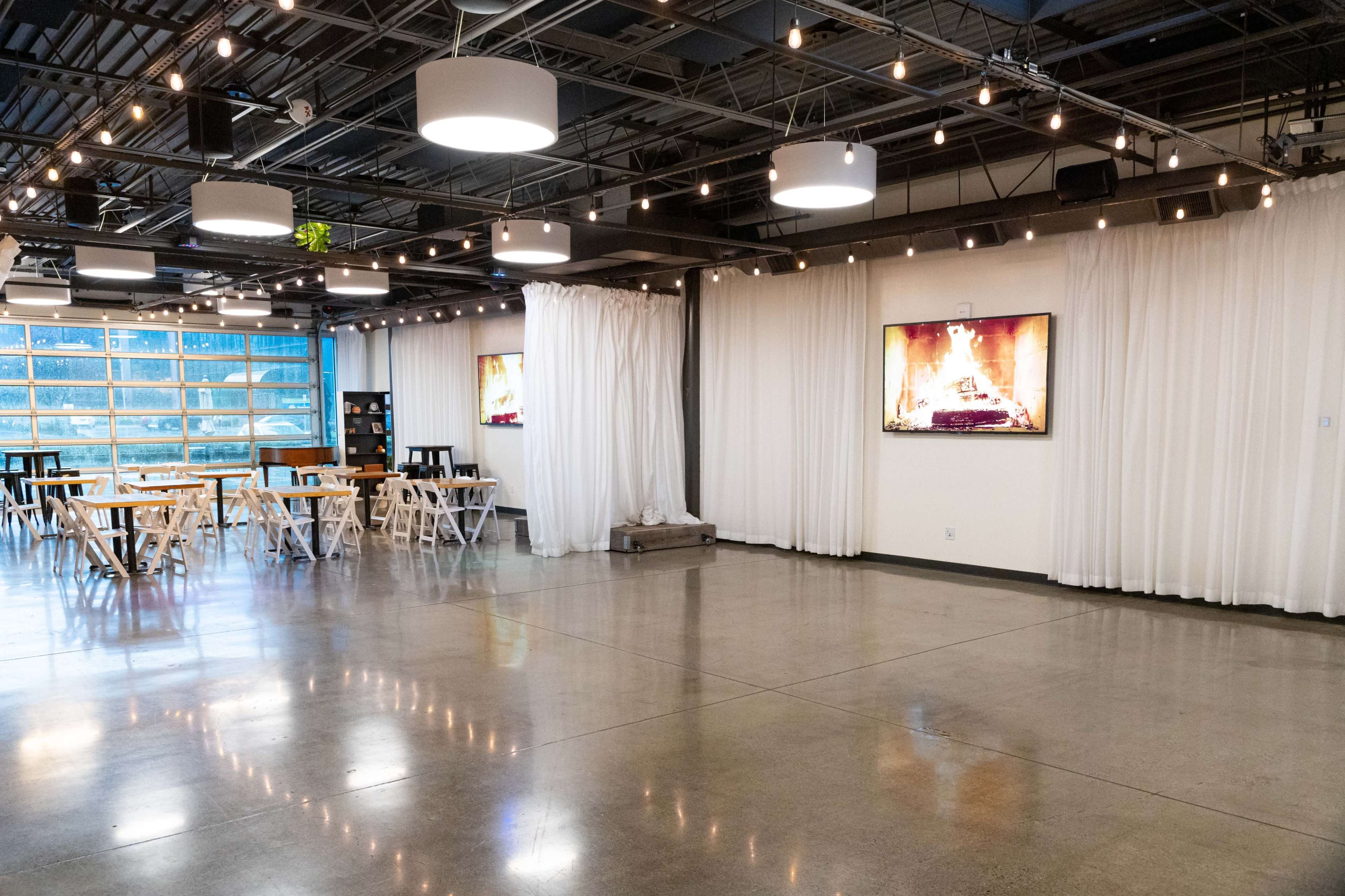 A spacious event room with white curtains, string lights, wooden tables, and two screens displaying a fireplace.