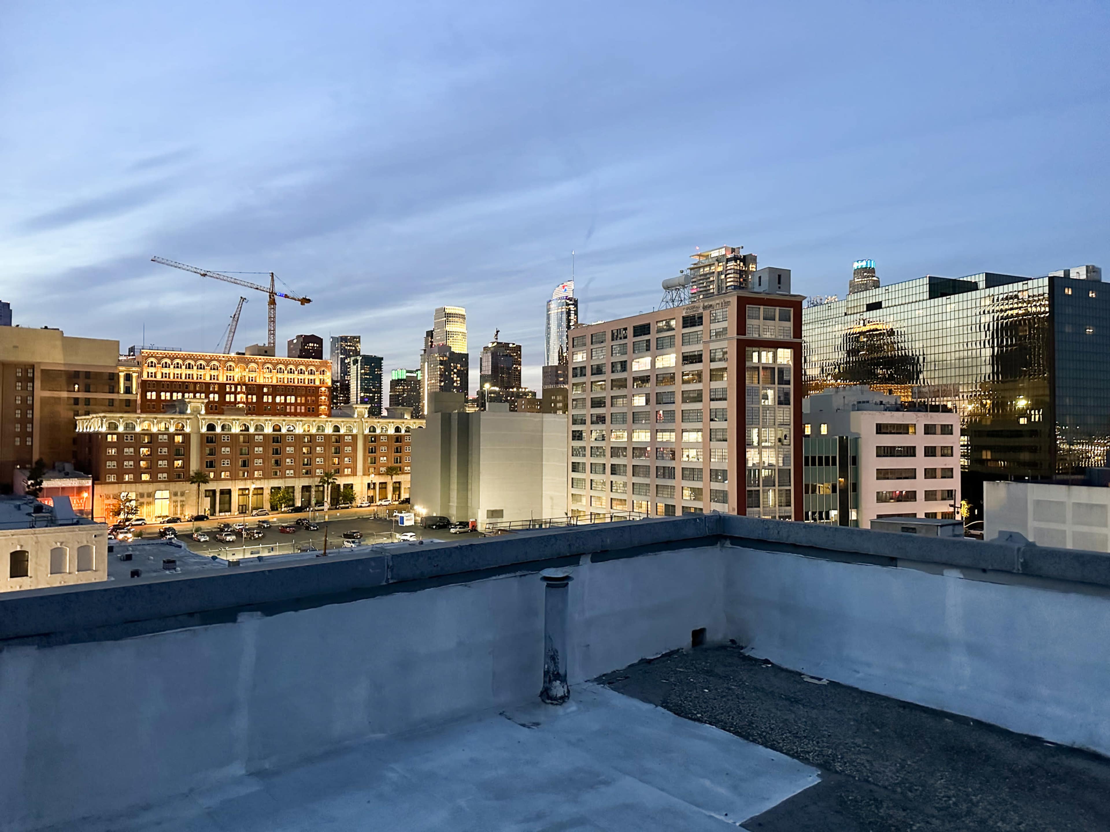 The image shows a rooftop view of a city skyline at dusk, with several buildings illuminated and construction cranes visible in the foreground.