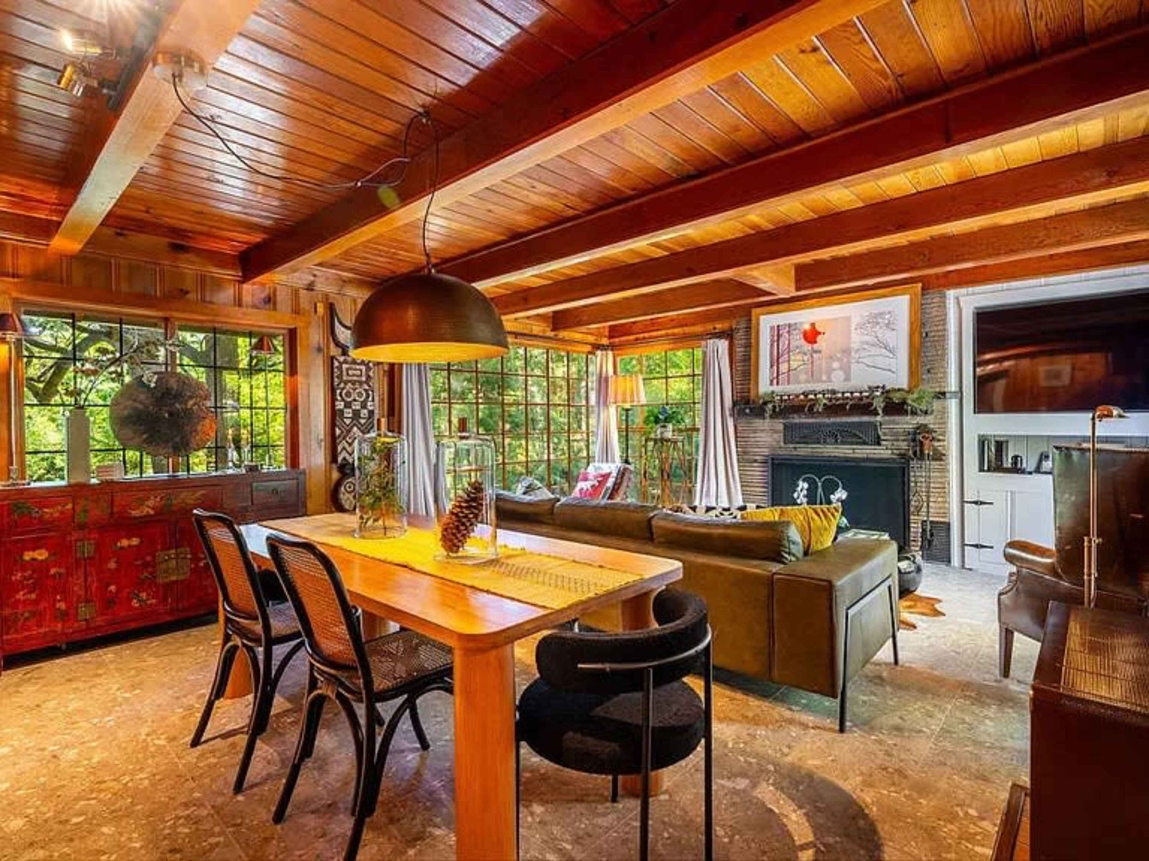 The image shows a cozy living room with wooden ceilings, a dining area, and large windows letting in natural light.