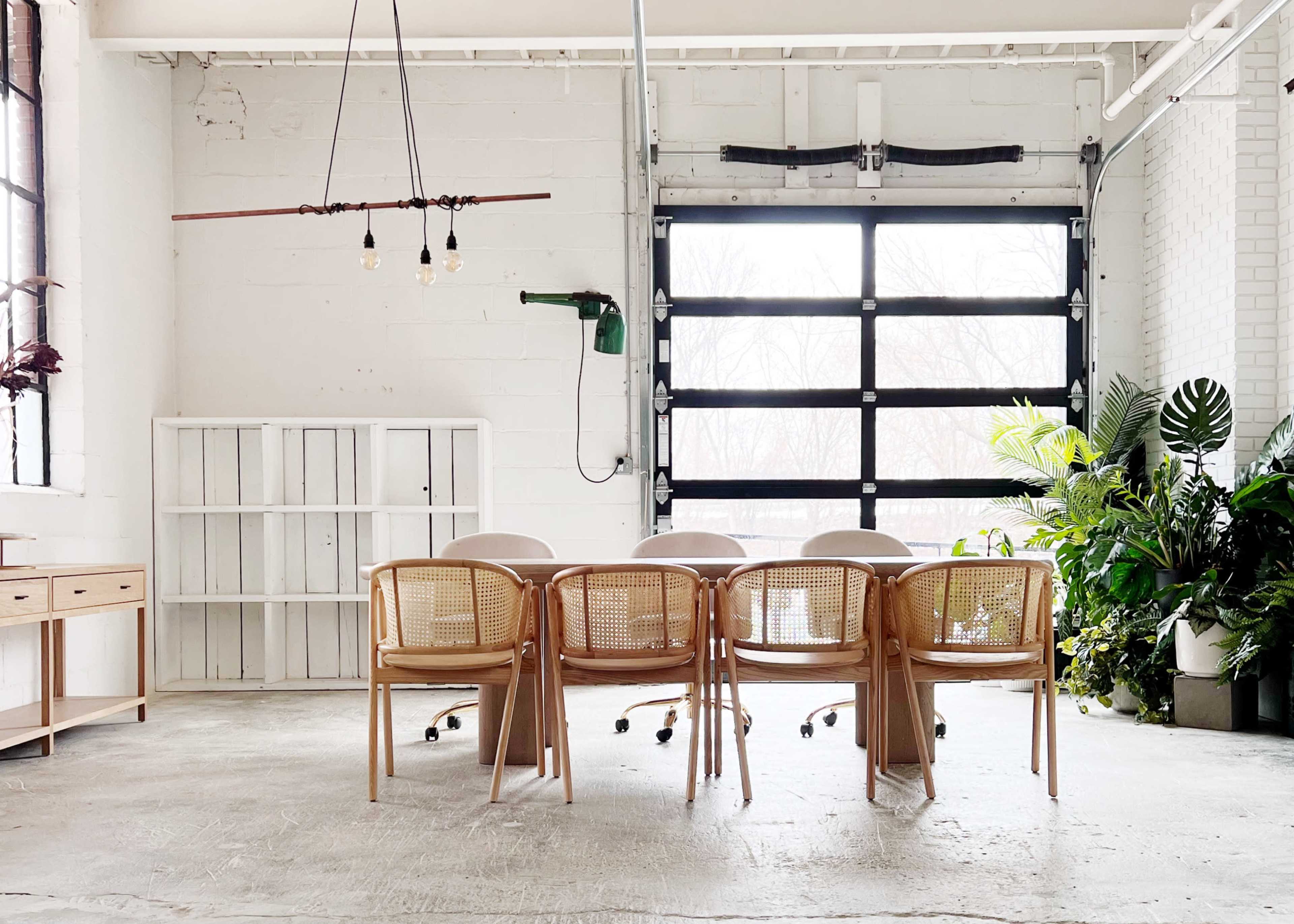 A modern meeting room features a long table surrounded by six chairs, with large windows and plants in the background.