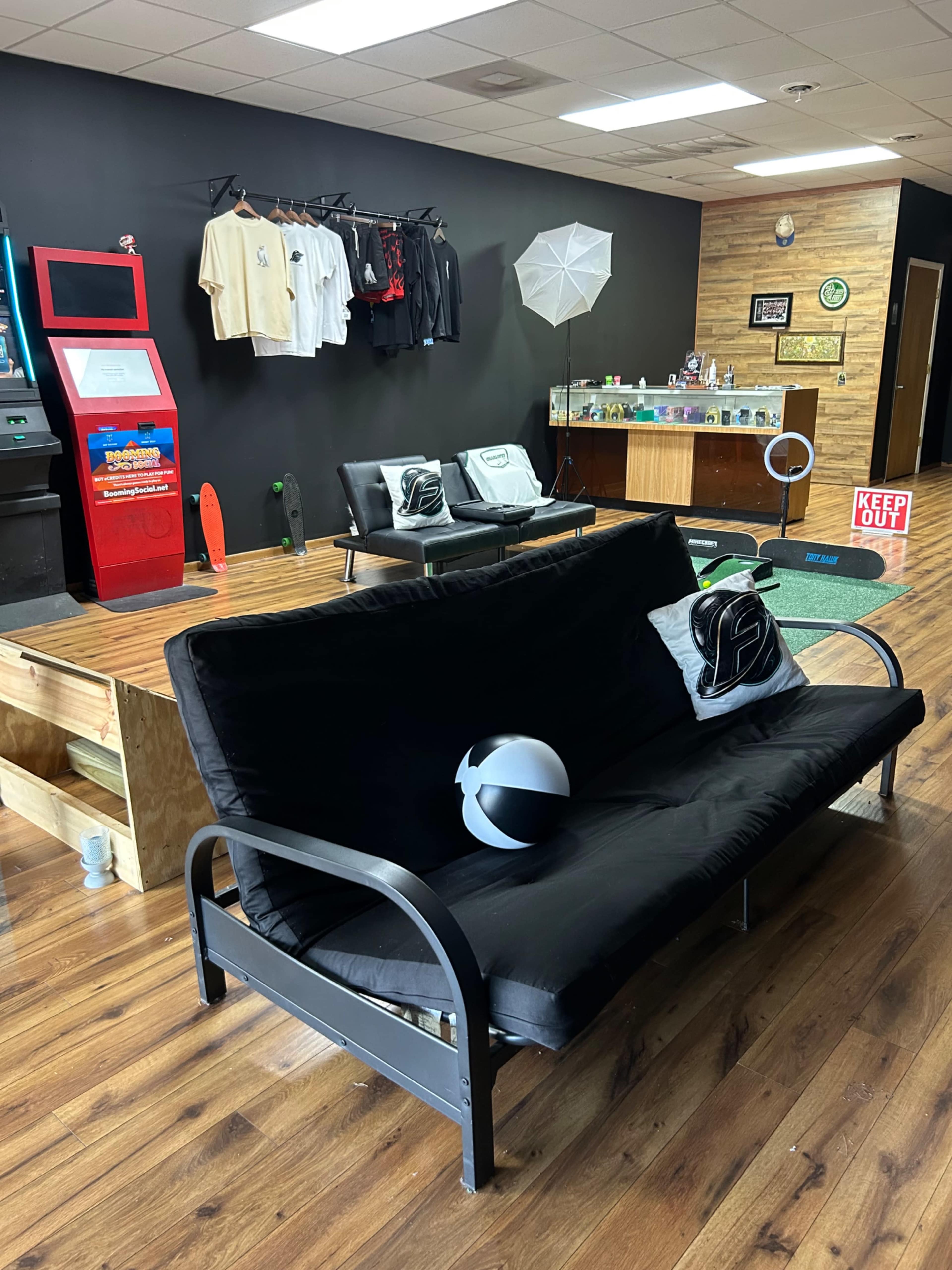 The image shows a modern lounge area with a black futon, a small bar counter, and various clothing displayed on hangers against a dark wall.