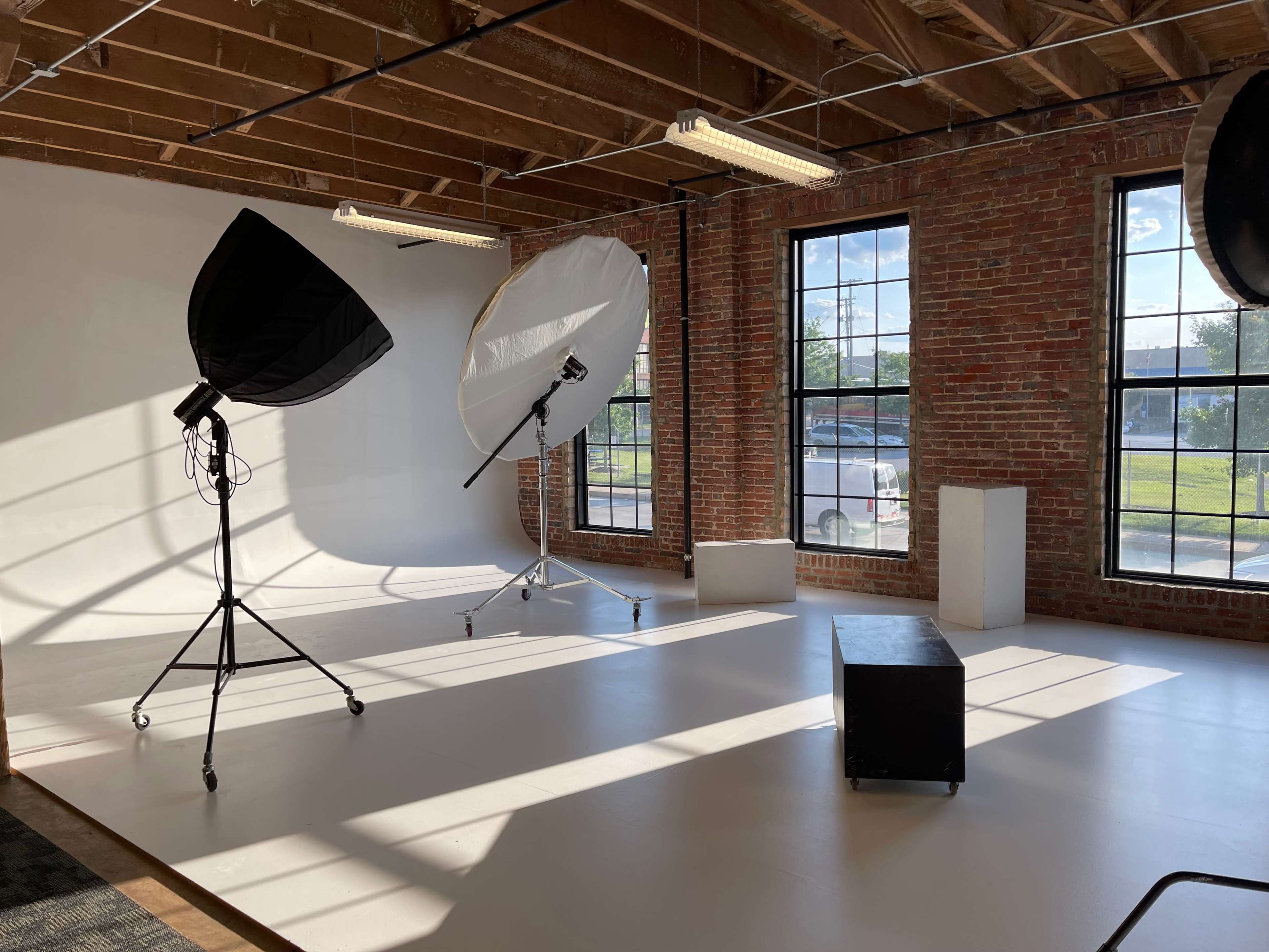 A spacious studio with large windows, brick walls, and photography lighting equipment set up on a white floor.