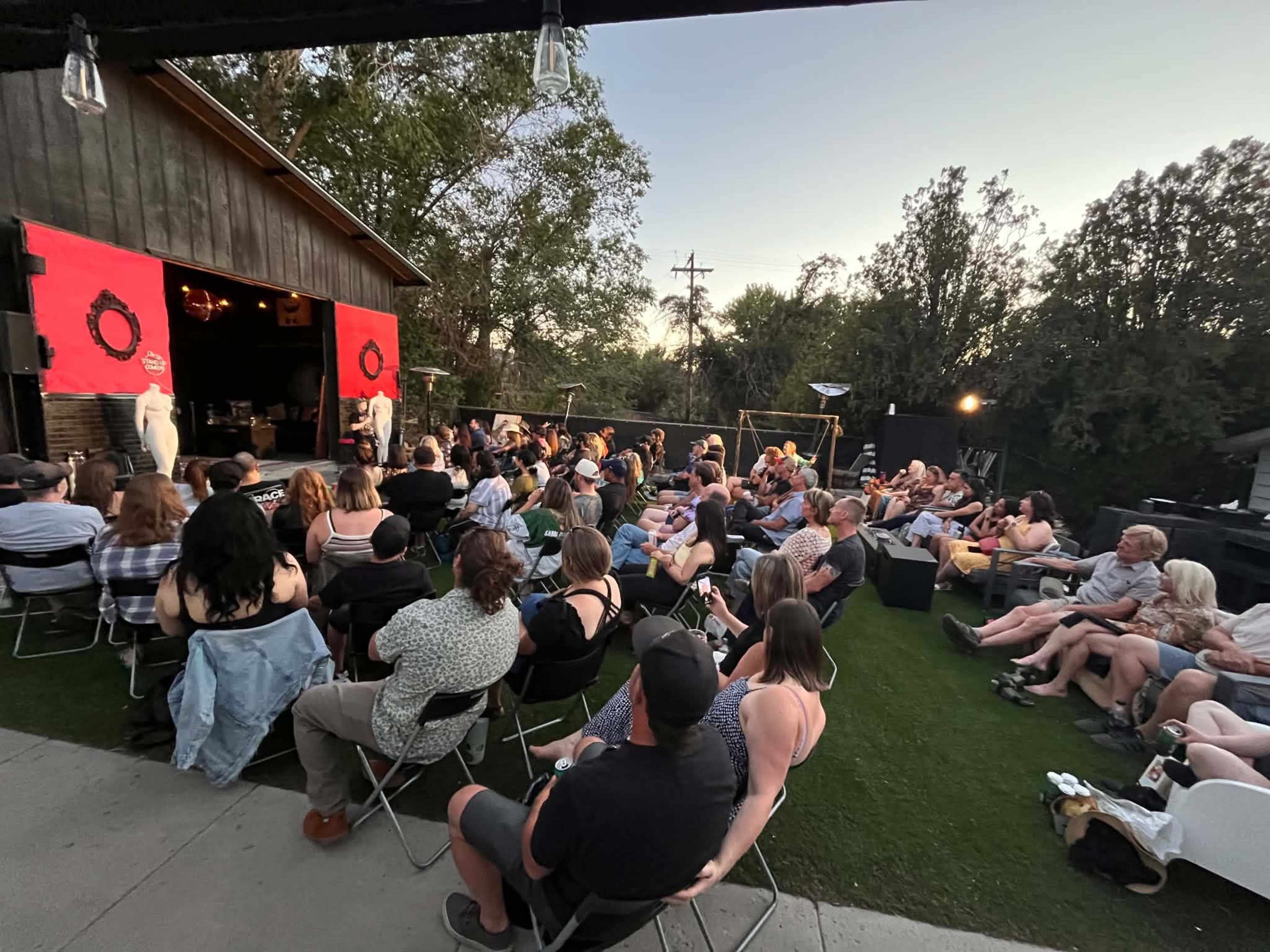 A large audience sits on chairs in front of a stage with red curtains, surrounded by trees and outdoor lighting as the sun sets.