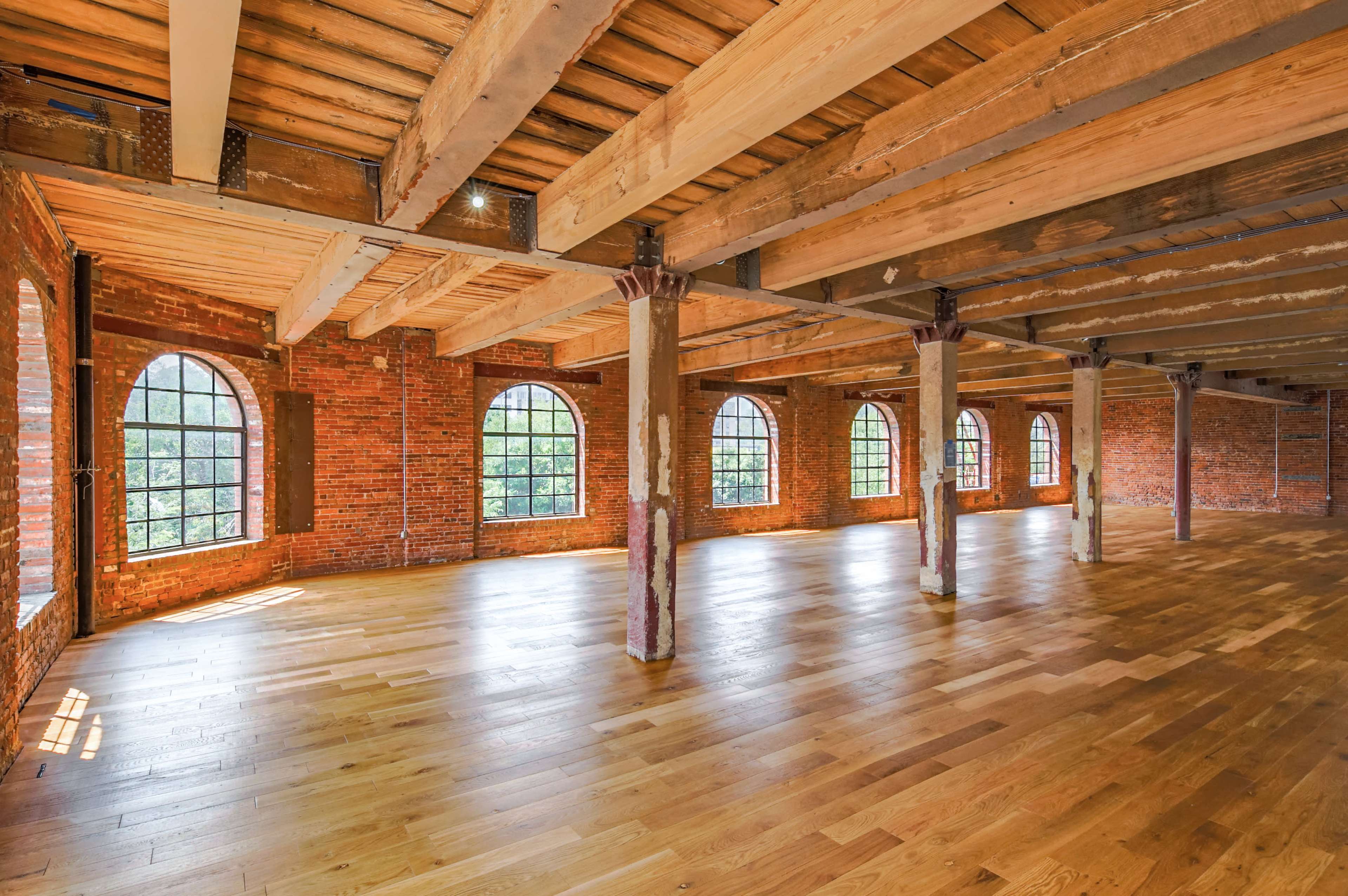 The image shows a spacious interior of a building with exposed brick walls, large windows, and wooden flooring.