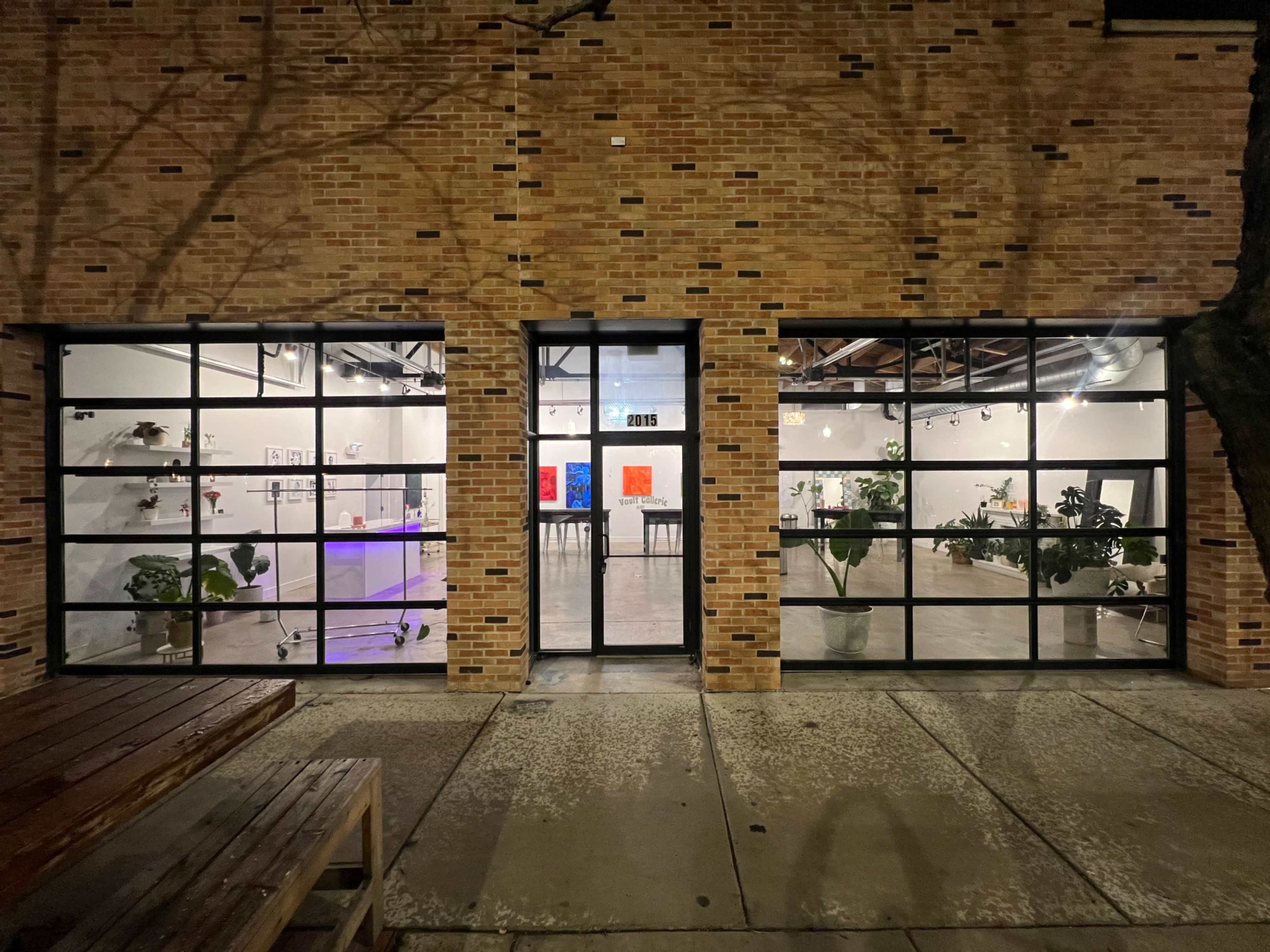 A brick building with large glass windows revealing a modern interior space featuring art displays and plants.