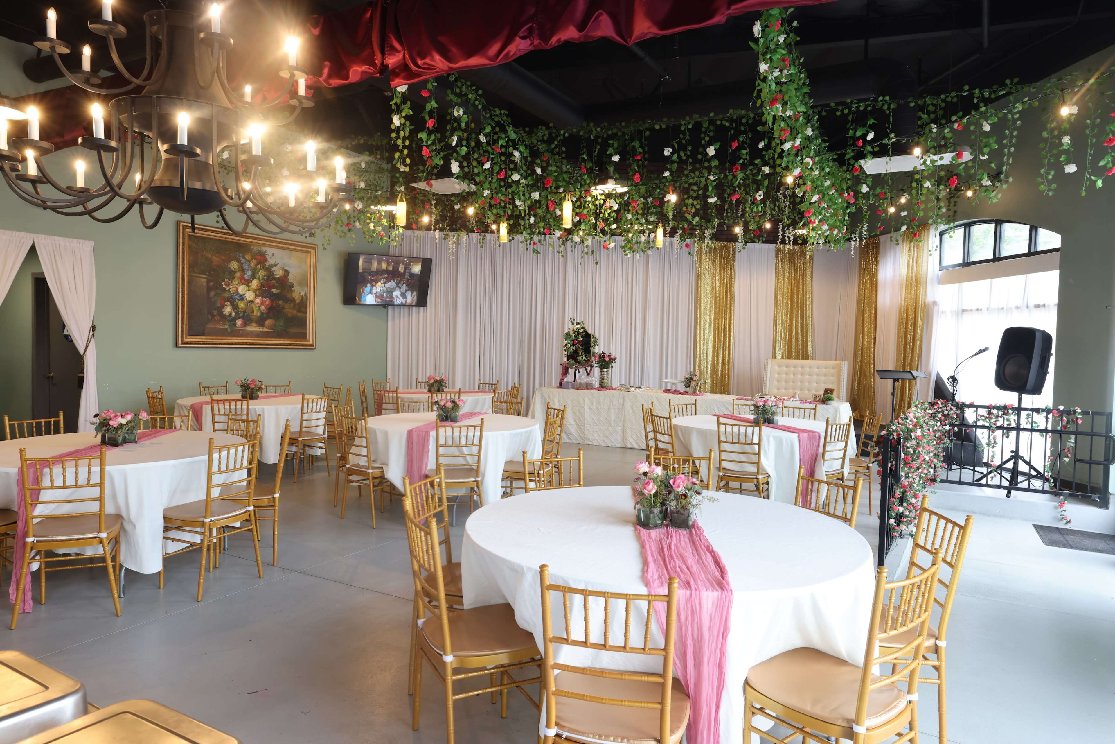 The image shows a decorated event space with round tables covered in white cloths, gold chairs, and floral arrangements, featuring a backdrop of draped fabric and greenery.