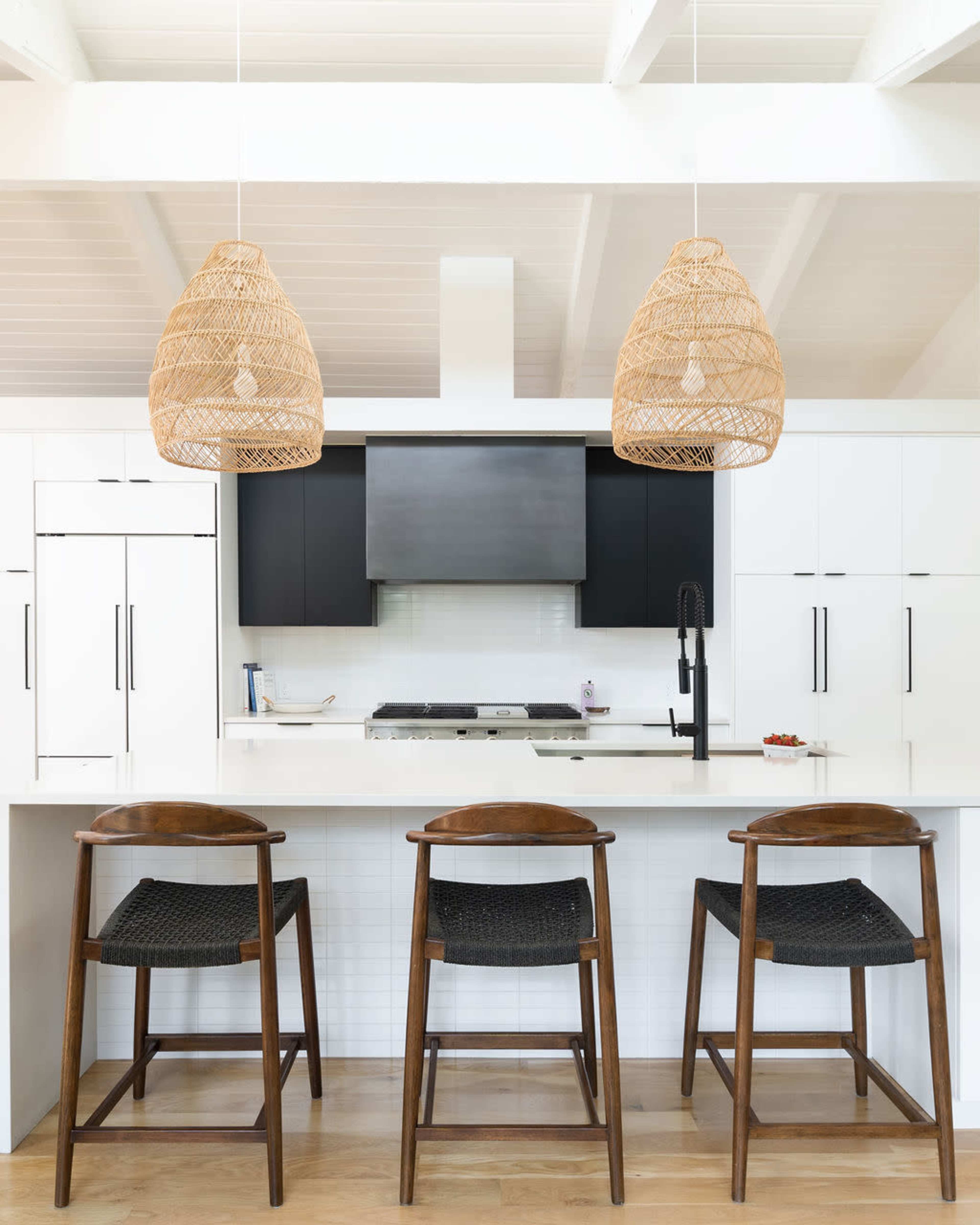 A modern kitchen features a central island with three wooden stools, white cabinetry, and two woven pendant lights hanging overhead.