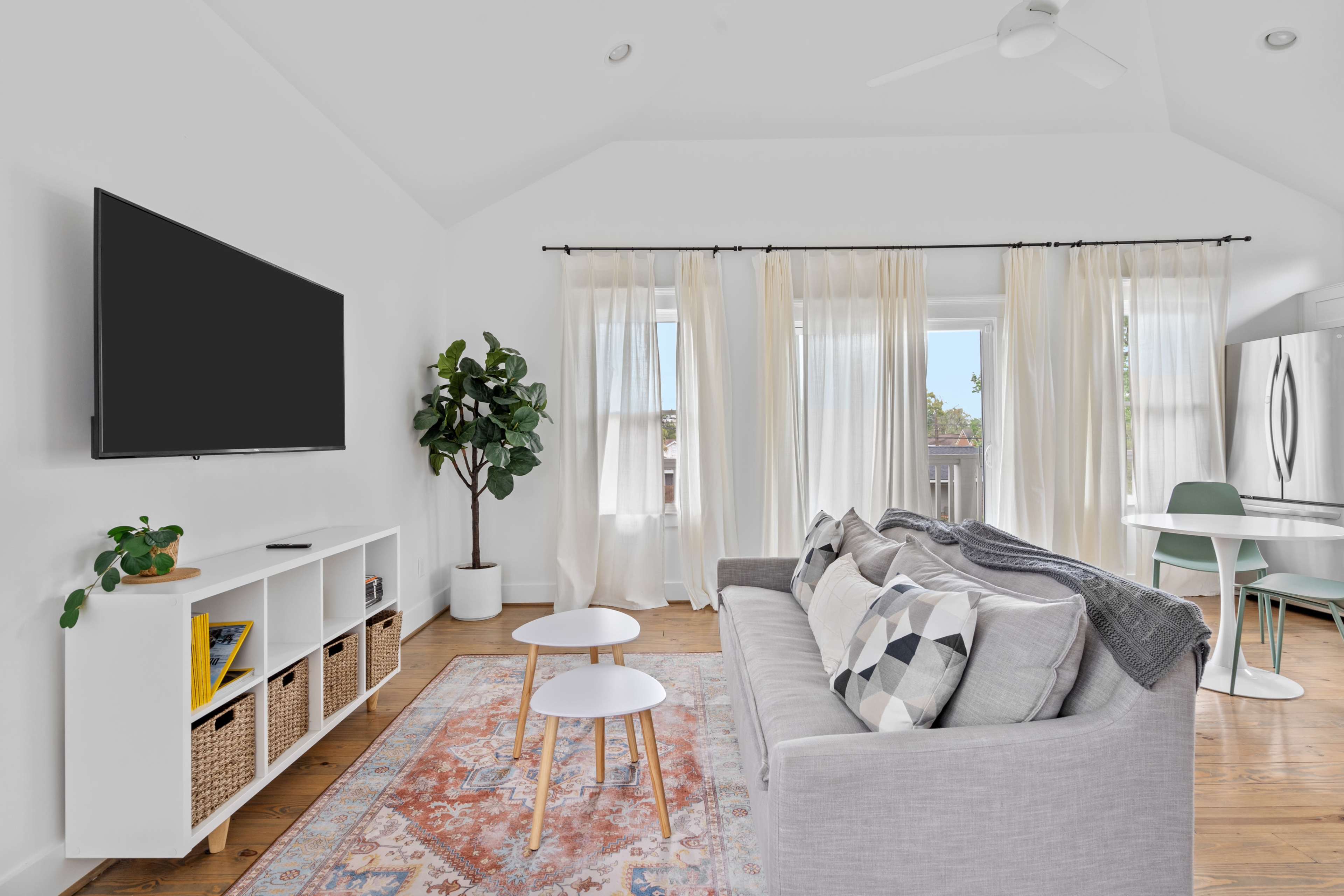 A modern living room features a gray sofa, a white TV stand, and two small coffee tables, with large windows draped in sheer curtains.
