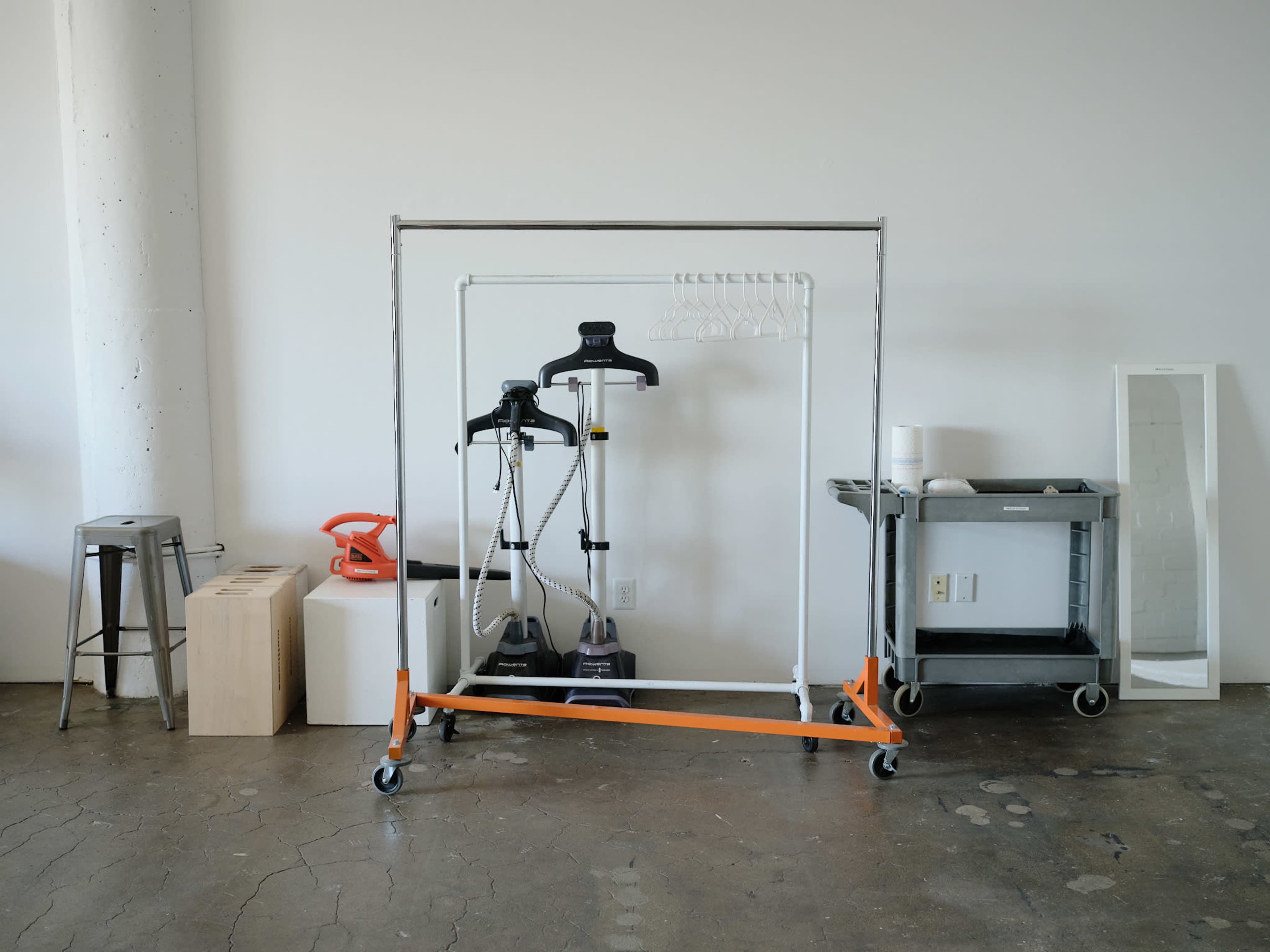 A clothing rack with two garment steamers, a mirror, a cart, a wooden box, and a stool against a plain wall.