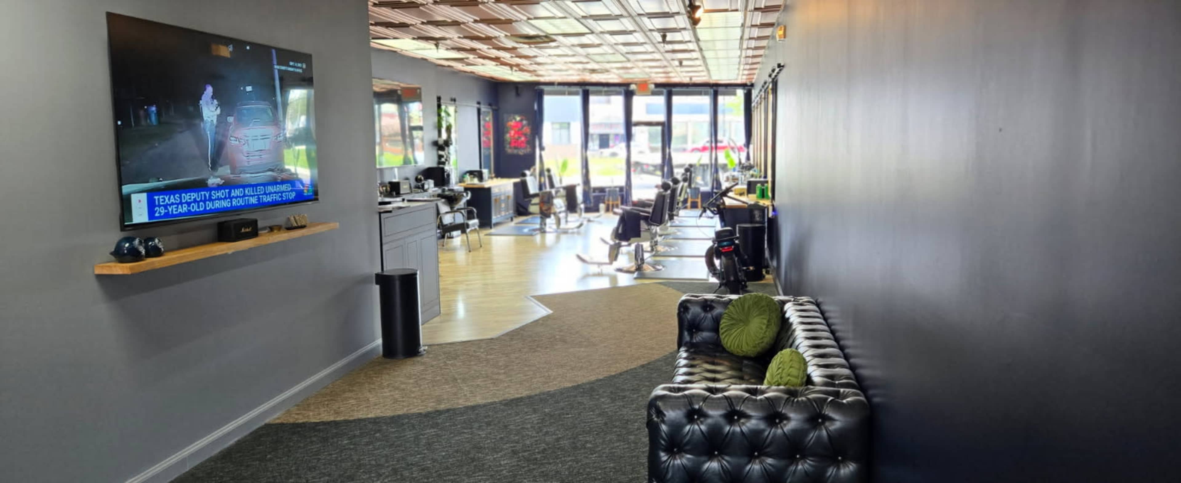 The image shows a modern salon interior featuring a black tufted sofa, a large television displaying news, and several salon chairs along the walls.
