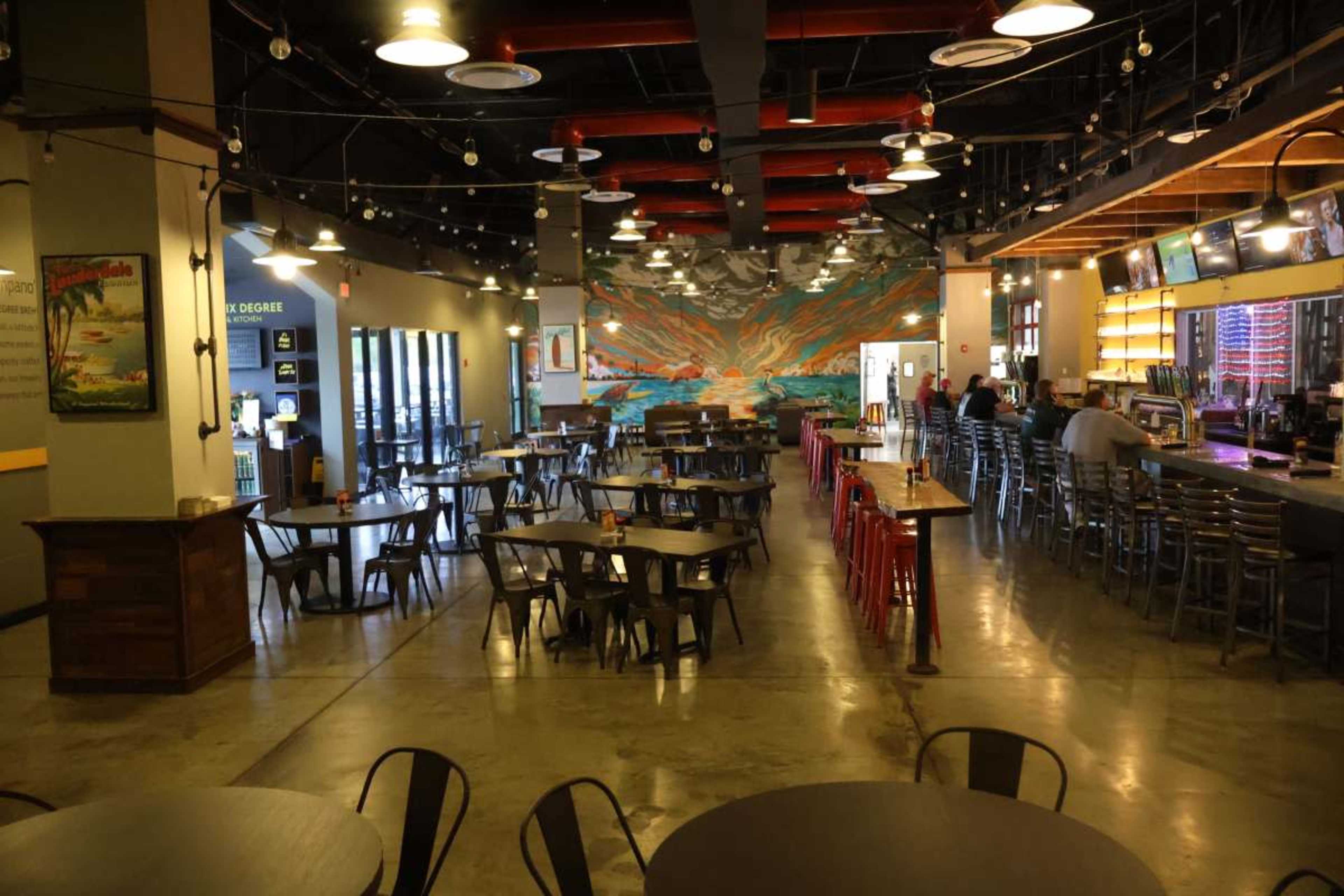 The image shows a spacious restaurant interior with a colorful mural, metal tables, and chairs, and a bar area at one end.