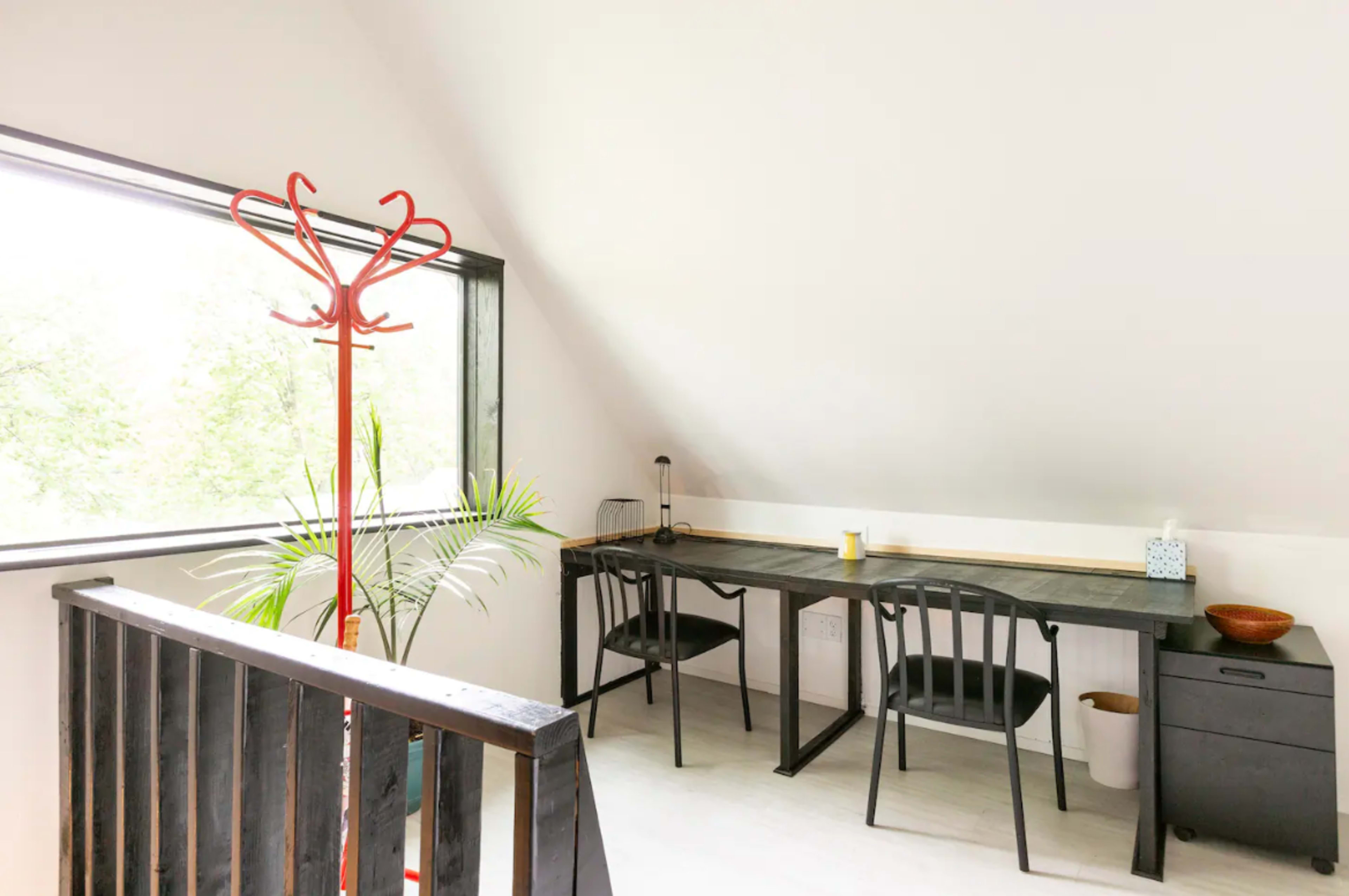 A minimalist workspace features a long black desk, two chairs, and a red coat rack, with large windows overlooking greenery.