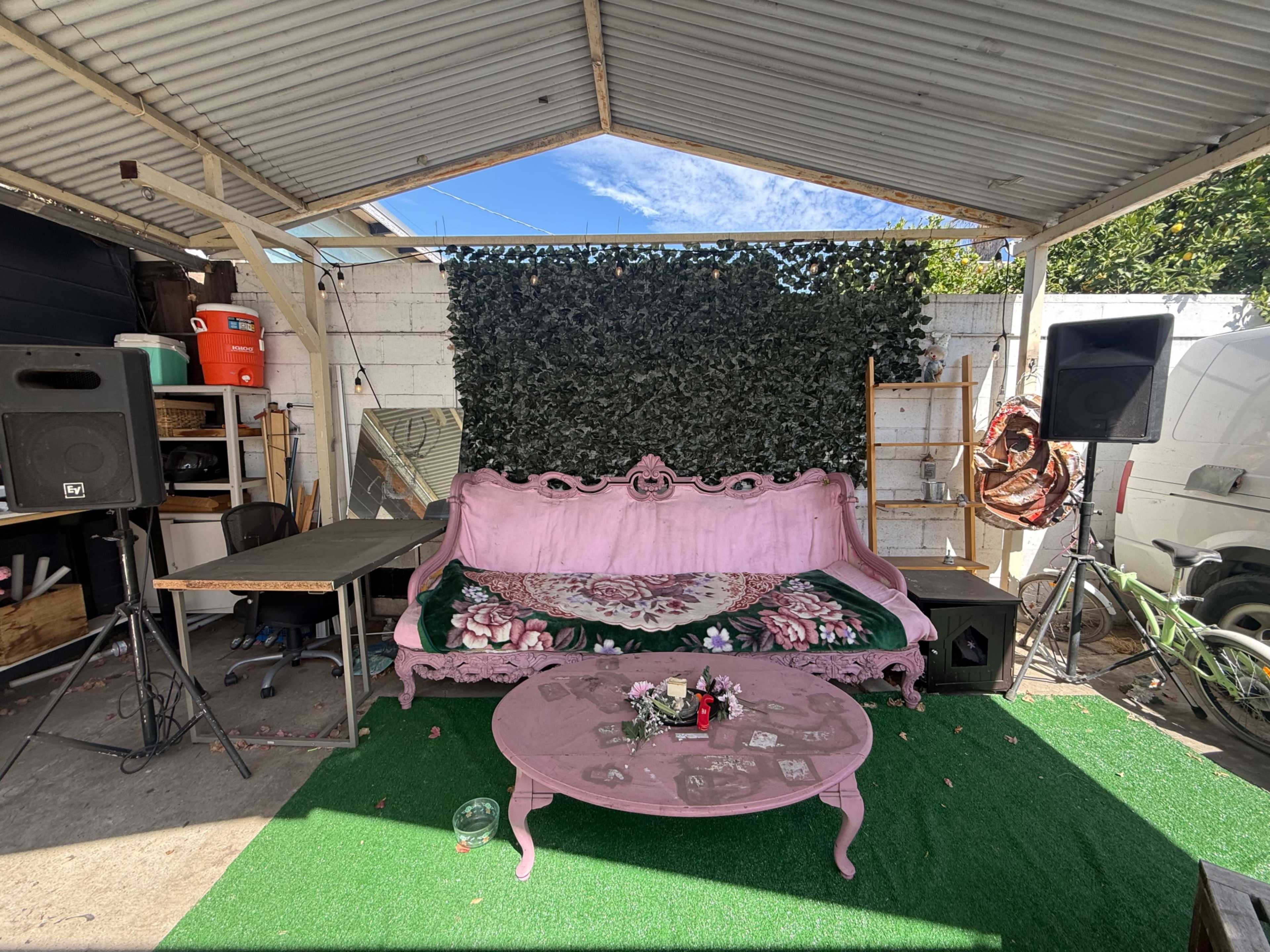 The image shows a covered outdoor space featuring a pink vintage sofa, a green carpet, a coffee table with decorations, and speakers on either side.