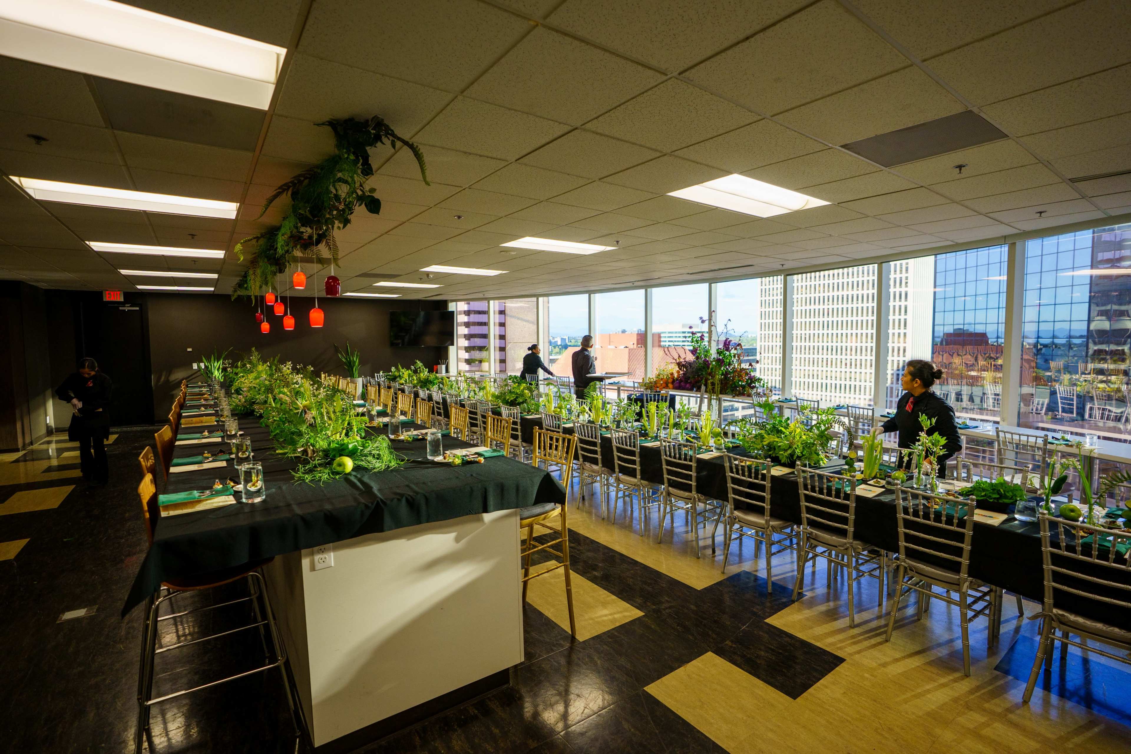 A long dining table is set with greenery and tableware, while staff prepare the room with large windows revealing a city skyline.
