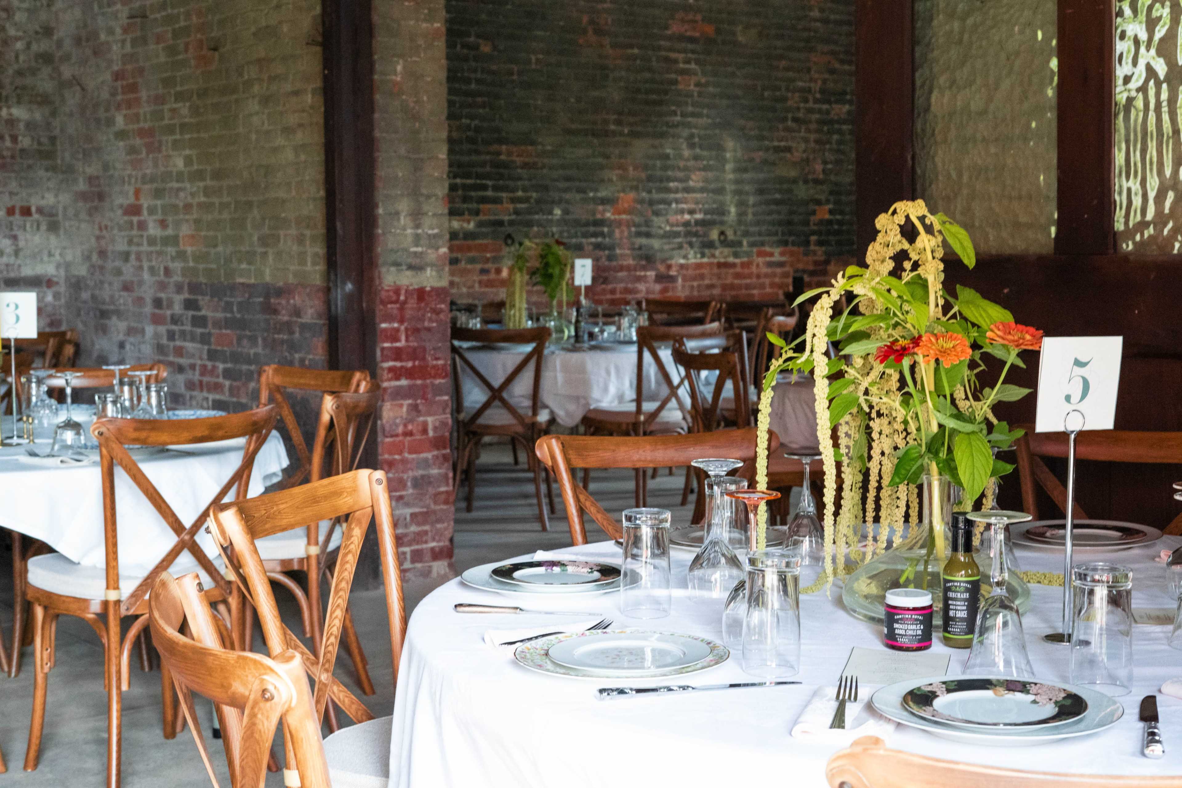 The scene features a dining area with wooden tables set for a meal, adorned with glassware and floral centerpieces.