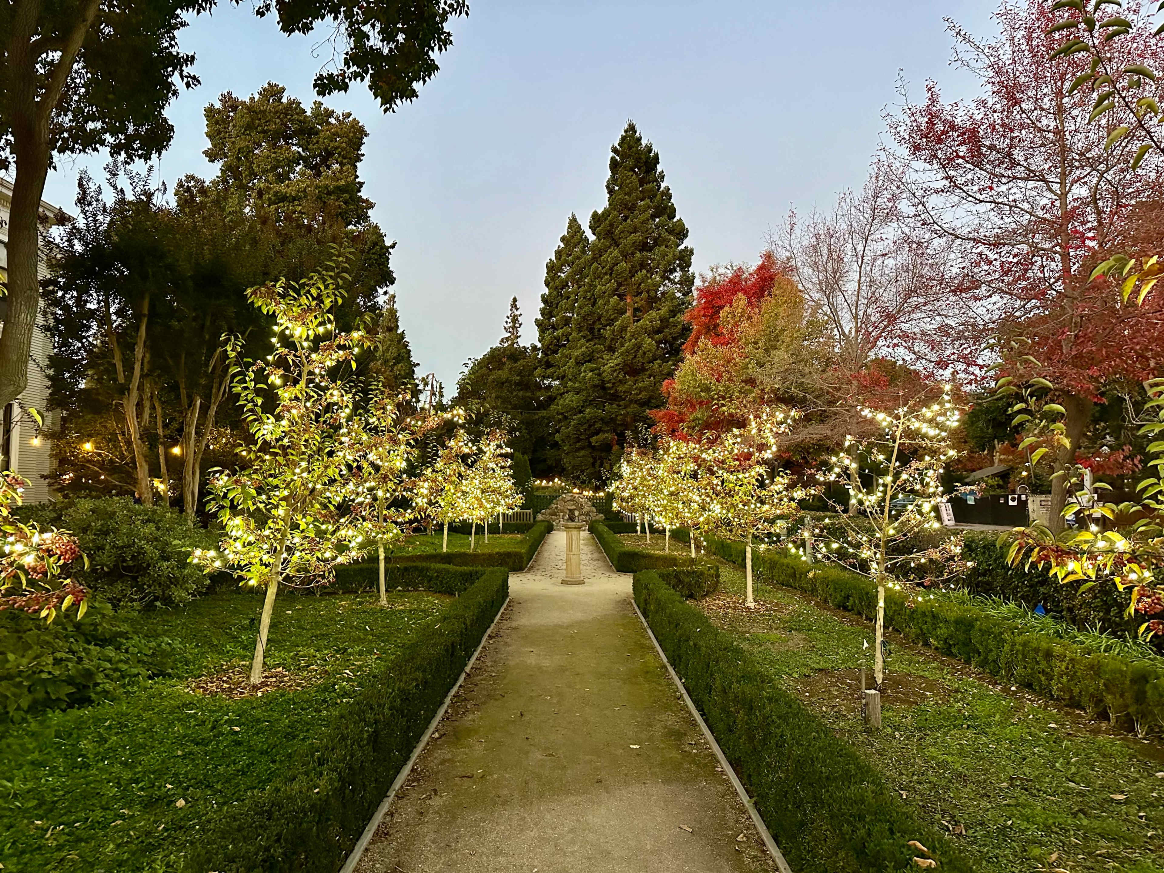 The scene depicts a landscaped garden pathway lined with trees adorned with lights, surrounded by neatly trimmed hedges and various foliage in an evening setting.