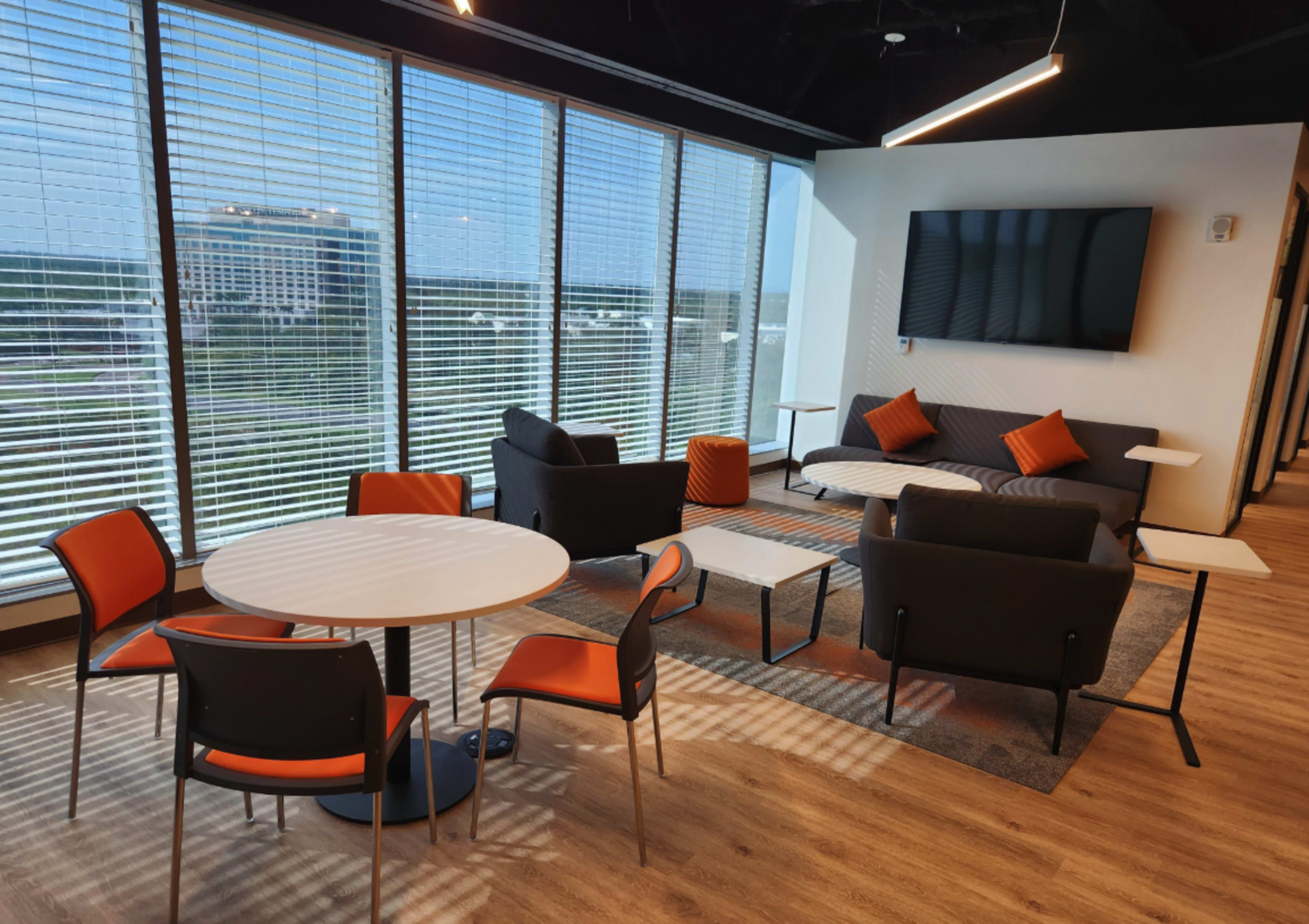 The image shows a modern lounge area with a sofa, armchairs, and tables, featuring large windows that provide a view of the outside.
