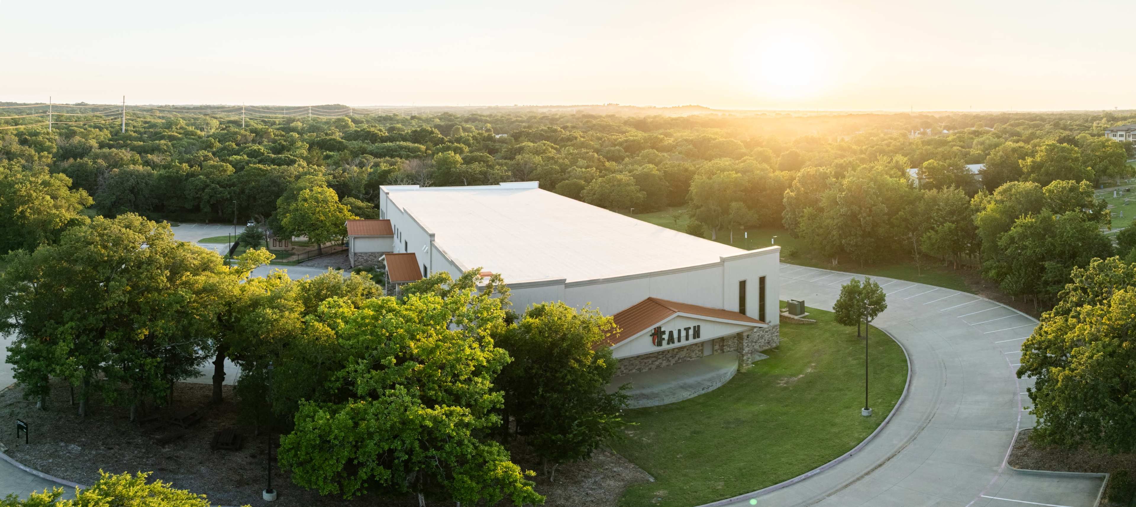 A large building with a flat roof is surrounded by trees and sits near a winding road, with the sun setting in the background.