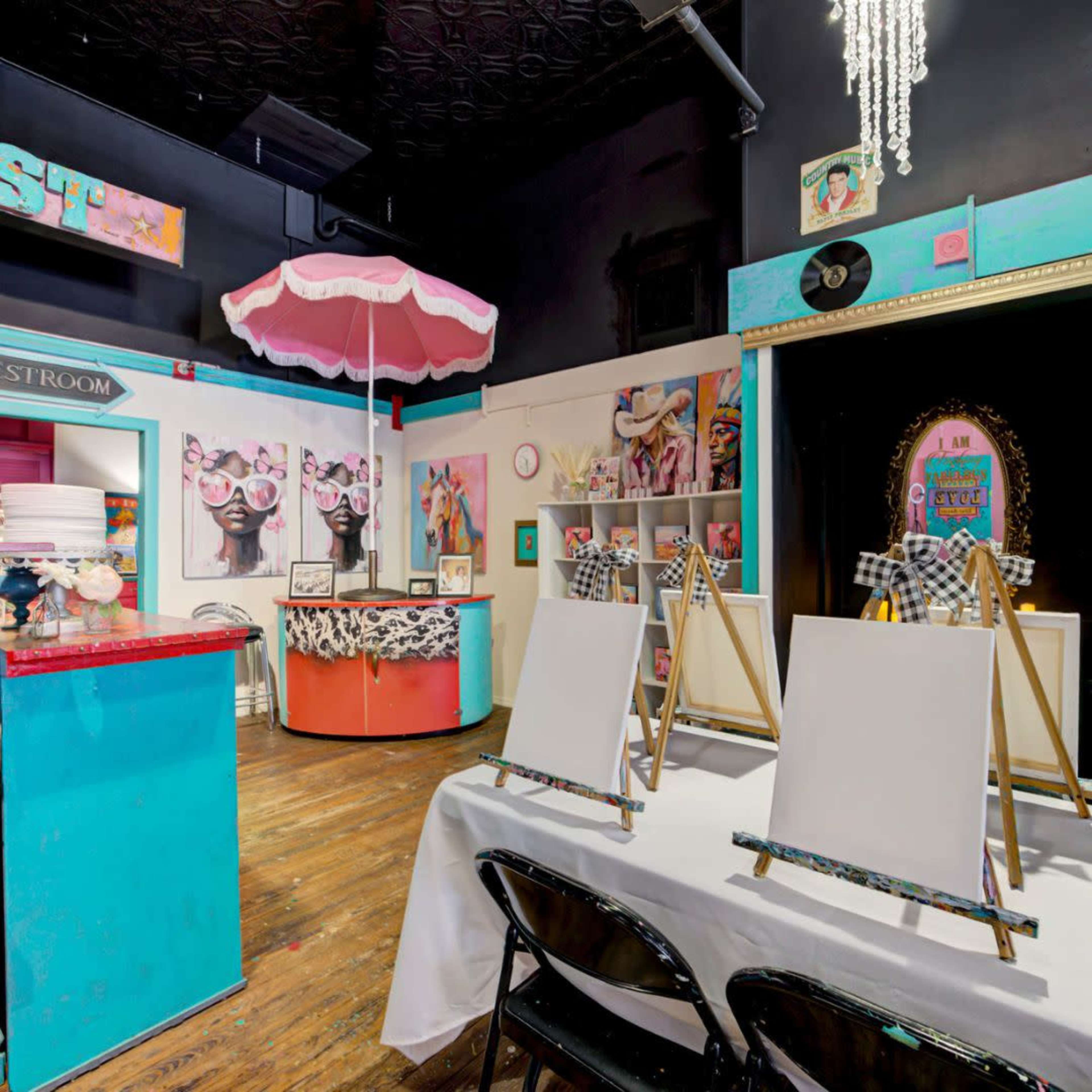 The image shows a colorful art studio featuring empty canvases on easels, a bar area, and decorative artwork on the walls.