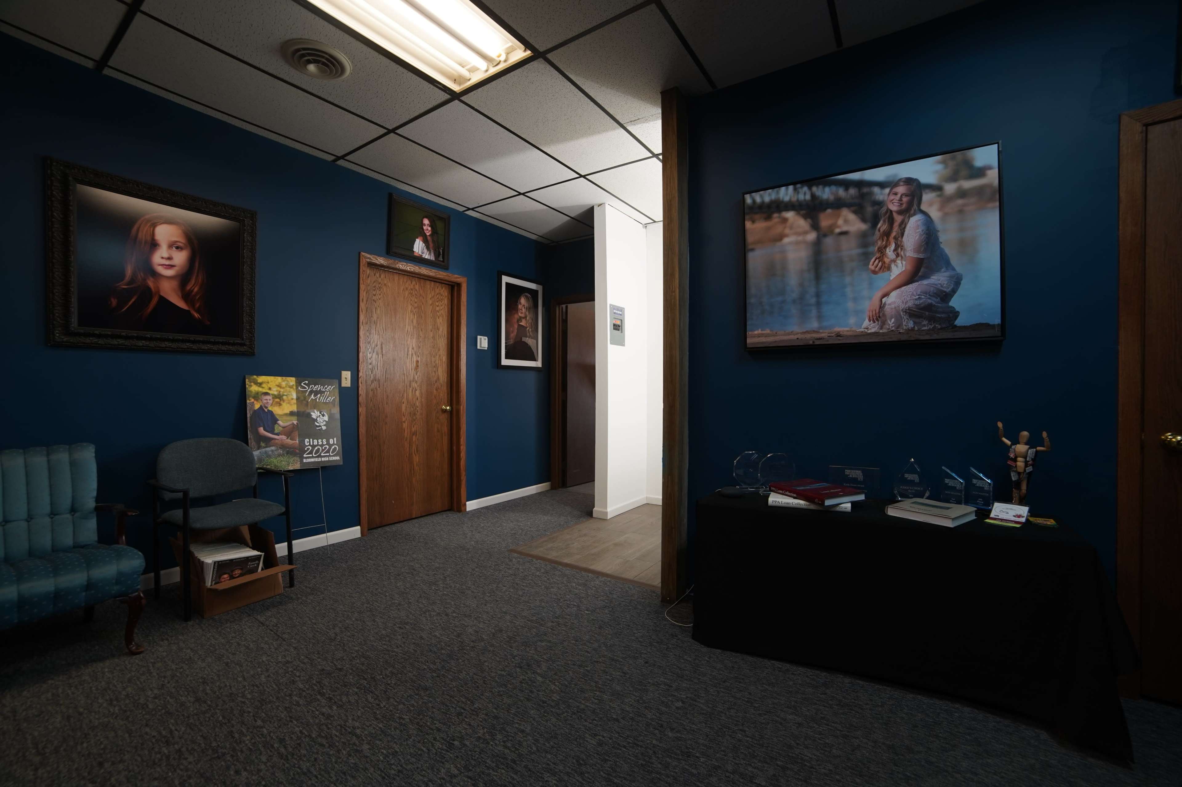The image shows a dimly lit room with dark blue walls featuring framed portraits, a seating area, and a hallway leading to other rooms.