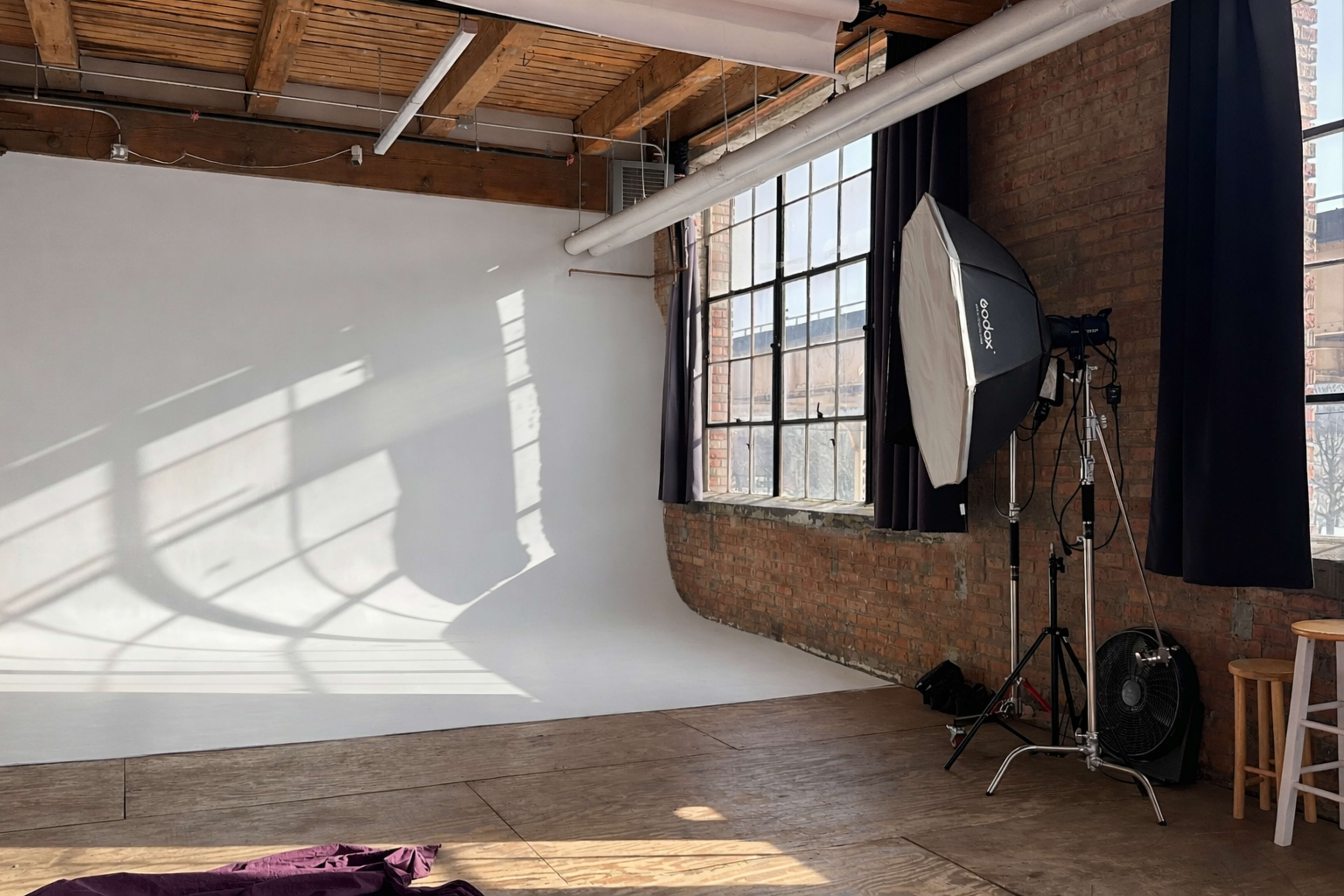 The image shows a photography studio with a white backdrop, softbox lighting, and large windows casting shadows on the wooden floor.