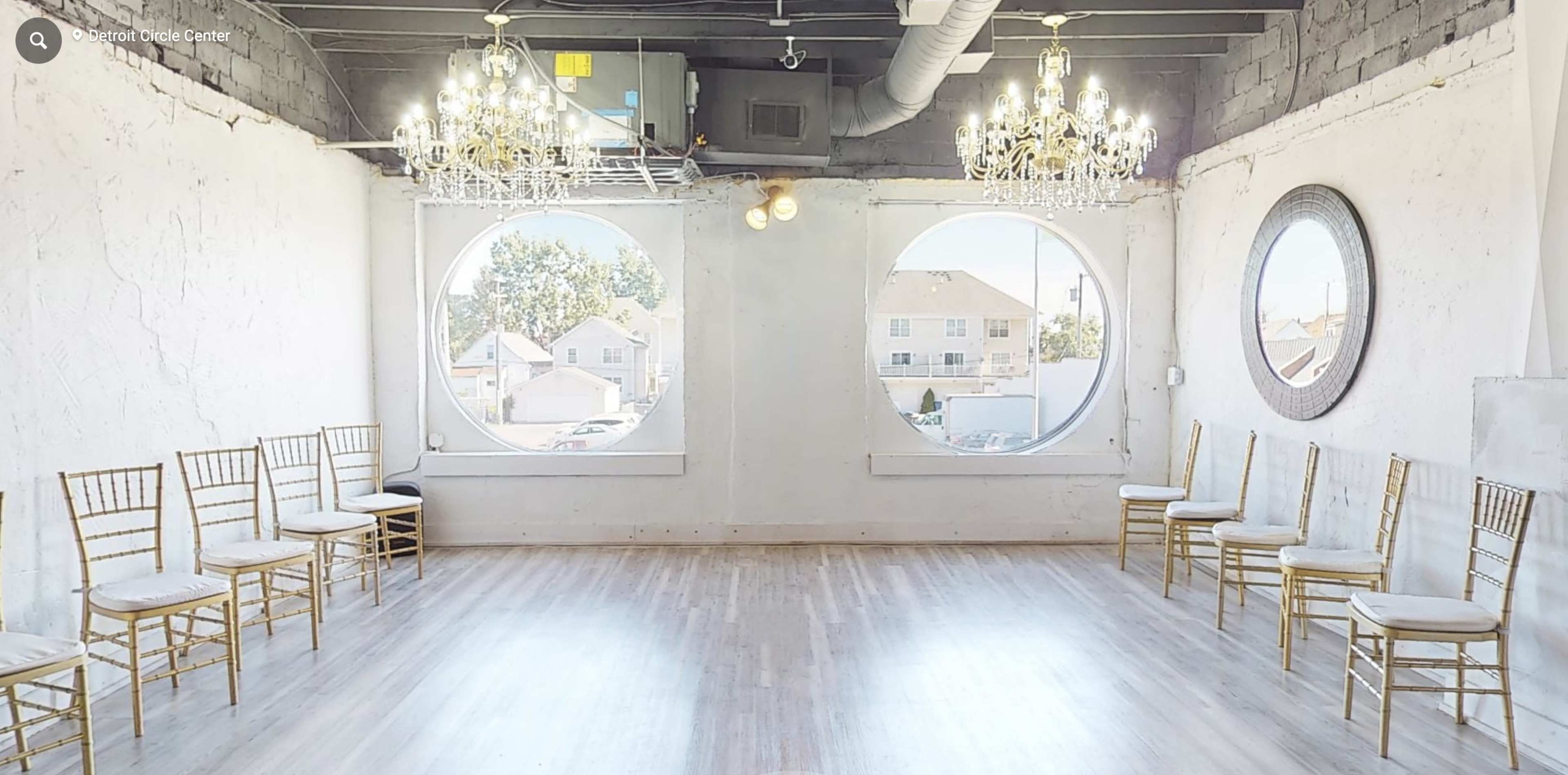 A spacious, well-lit room with two large circular windows, gold chandeliers, and a row of gold chairs along one wall.