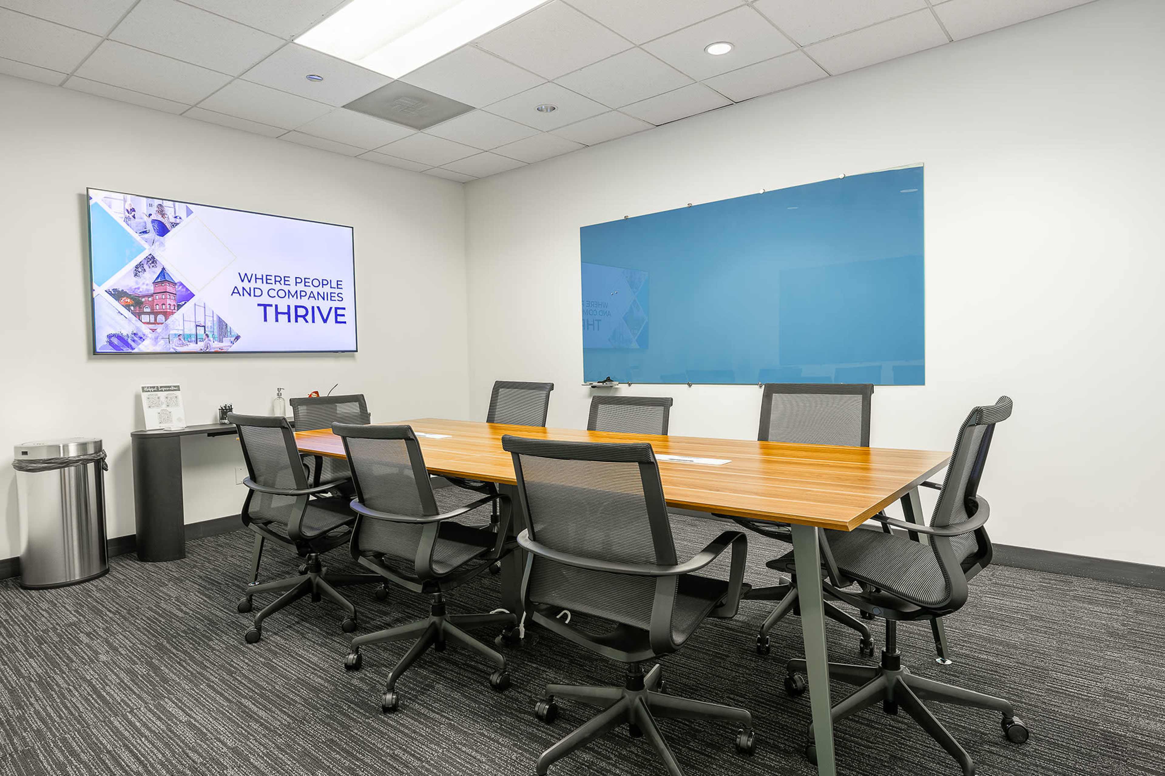The image shows a modern conference room with a rectangular wooden table surrounded by eight ergonomic chairs, a large screen displaying the word "THRIVE," and a whiteboard on the wall.