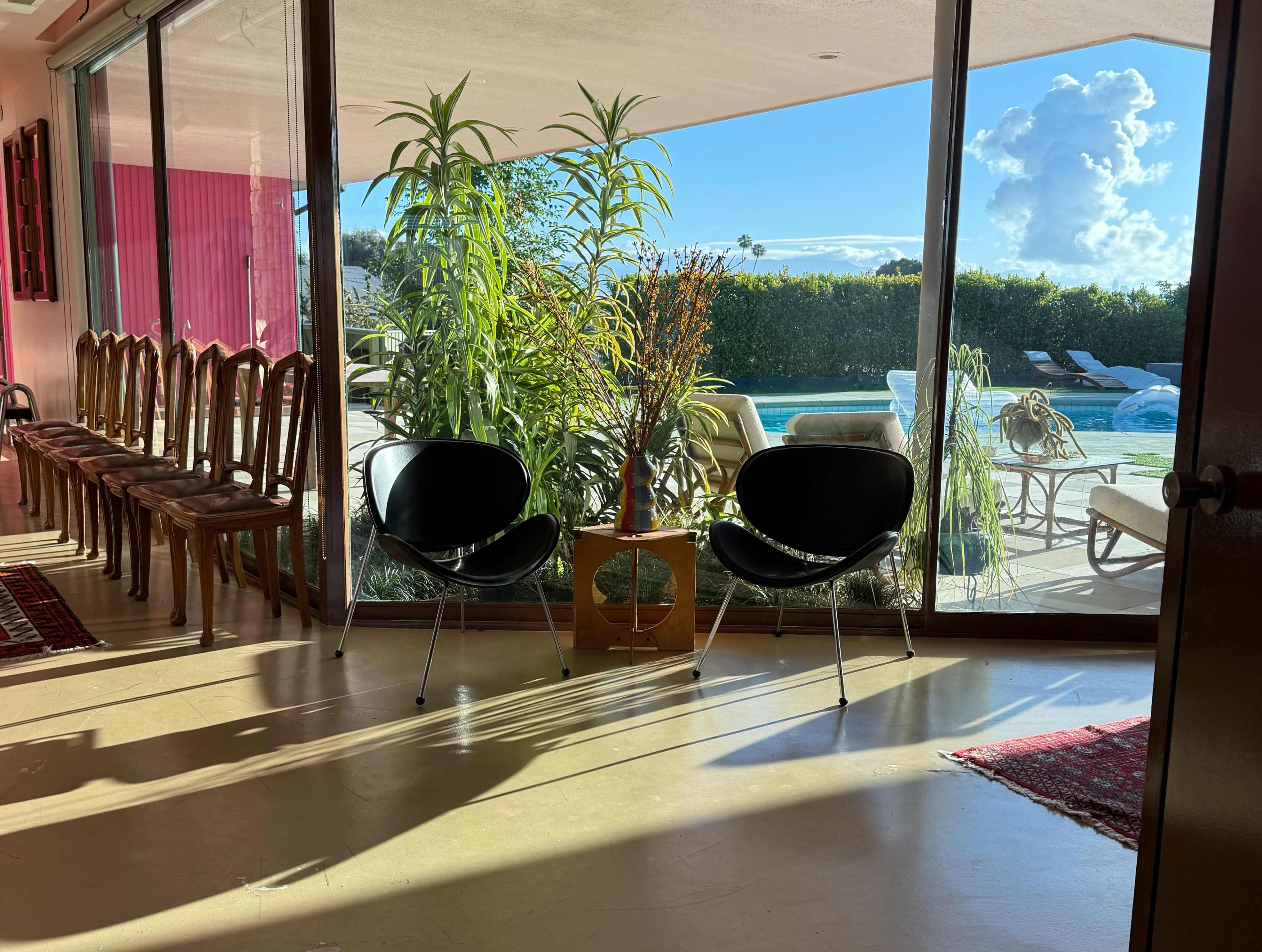 A pair of black chairs are positioned near a large window, showcasing a view of a swimming pool and a well-maintained garden outside.