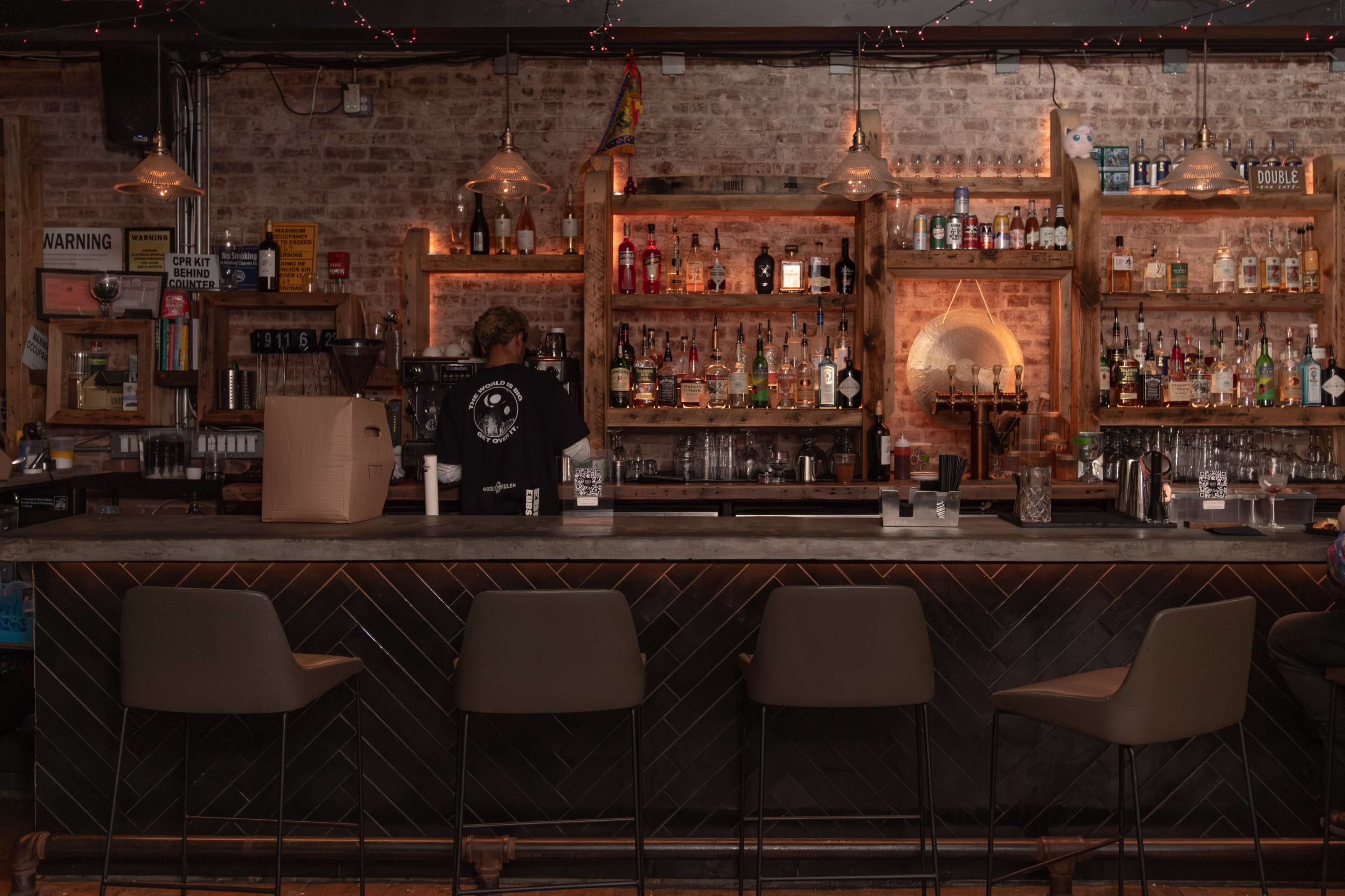 A bartender stands behind a well-stocked bar featuring shelves lined with various bottles of liquor and illuminated by pendant lights.