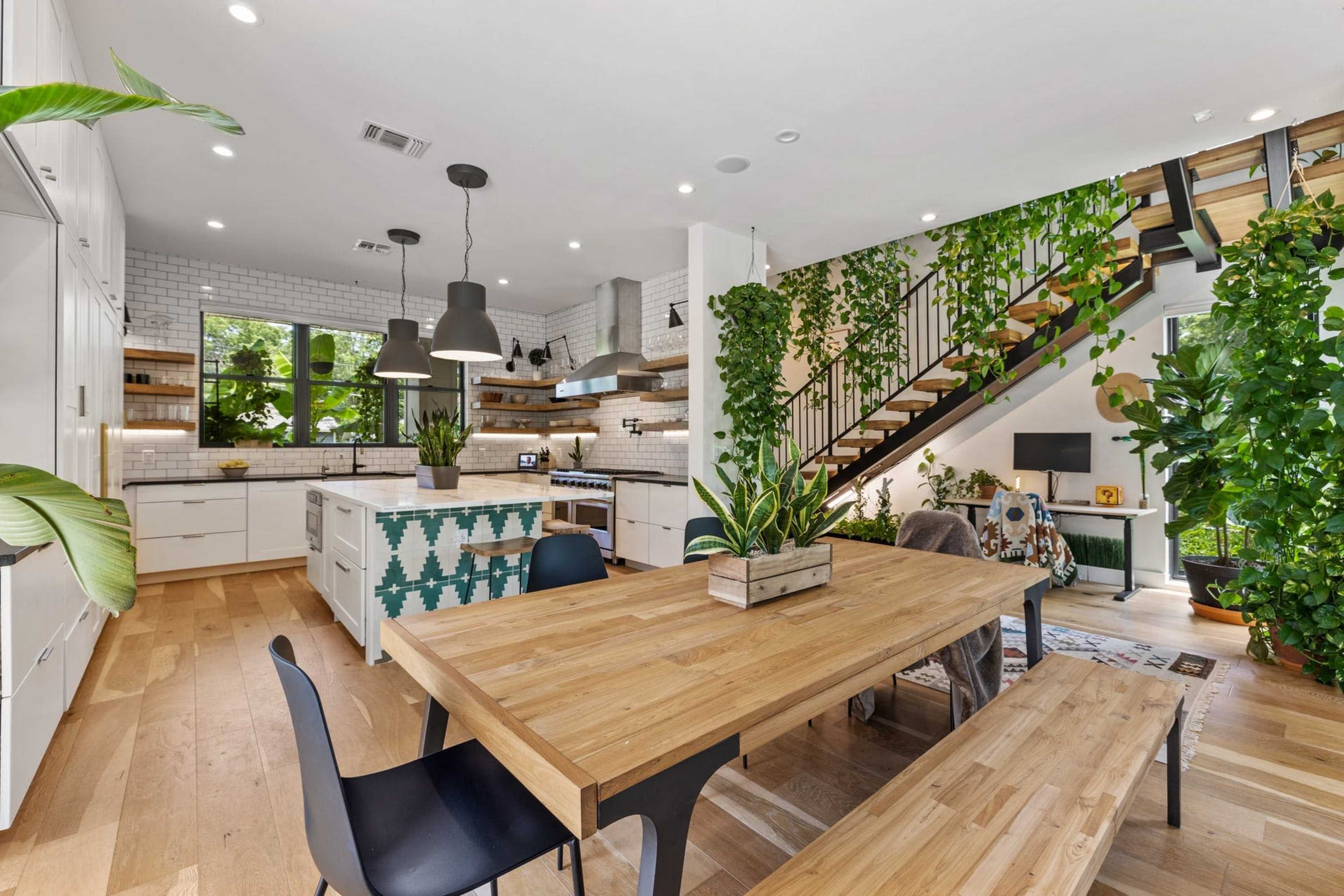 A modern kitchen and dining area features a wooden table, black chairs, and numerous plants, with a staircase in the background.