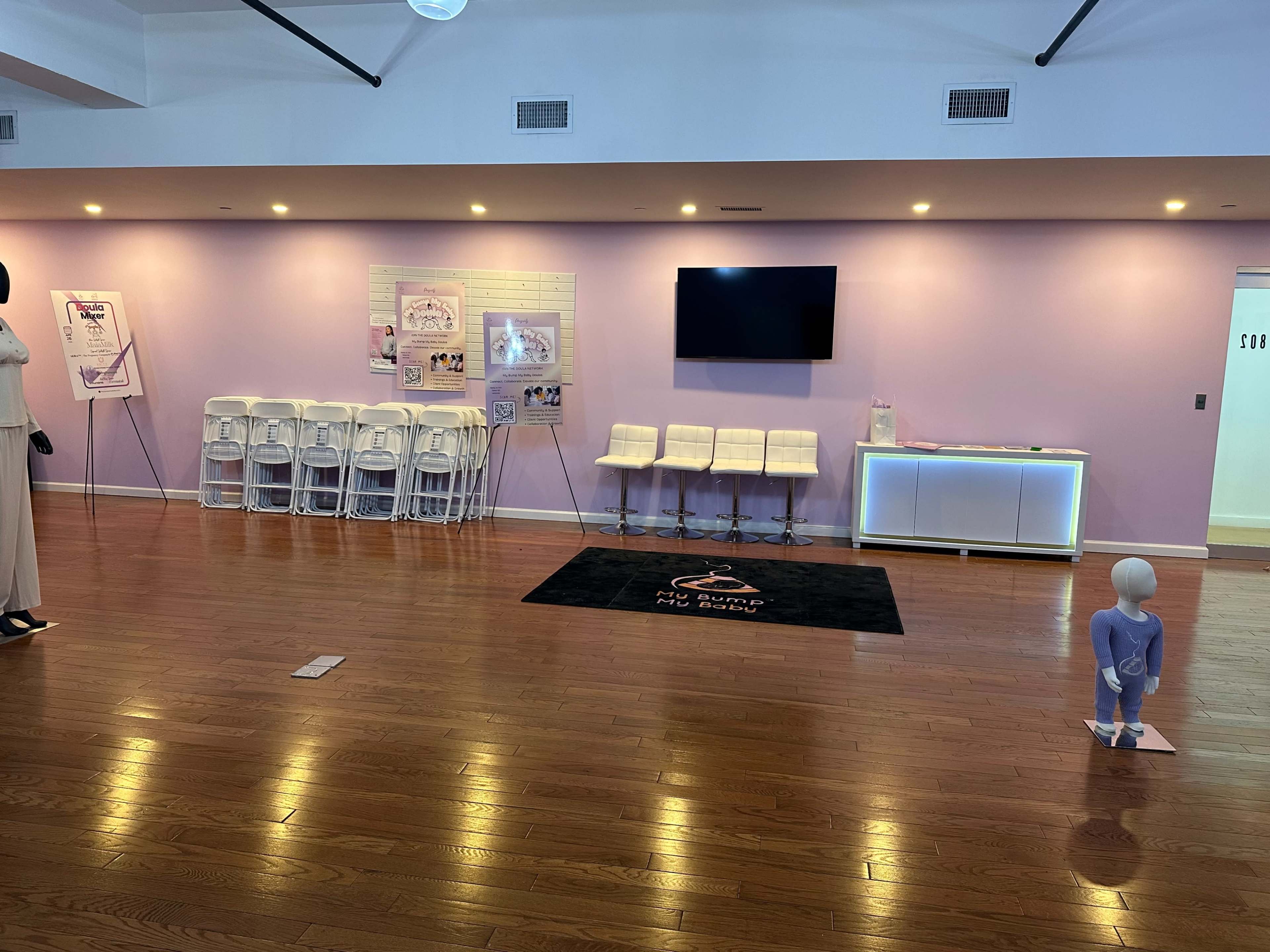 The image shows an empty room with hardwood flooring, purple walls, a television mounted on the wall, a row of white chairs, and a black rug with a logo.
