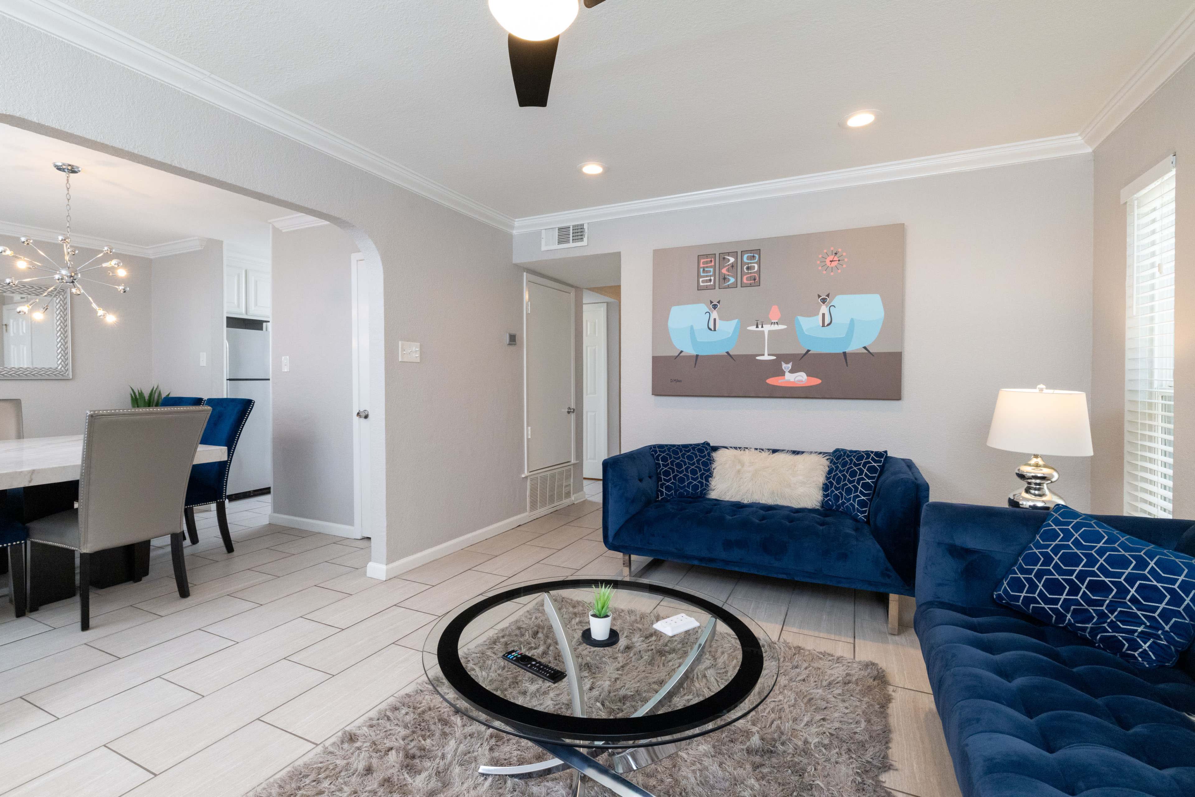 The image shows a modern living room with a blue couch, a glass coffee table, and a wall art piece featuring two animated characters, adjacent to a dining area with blue chairs.