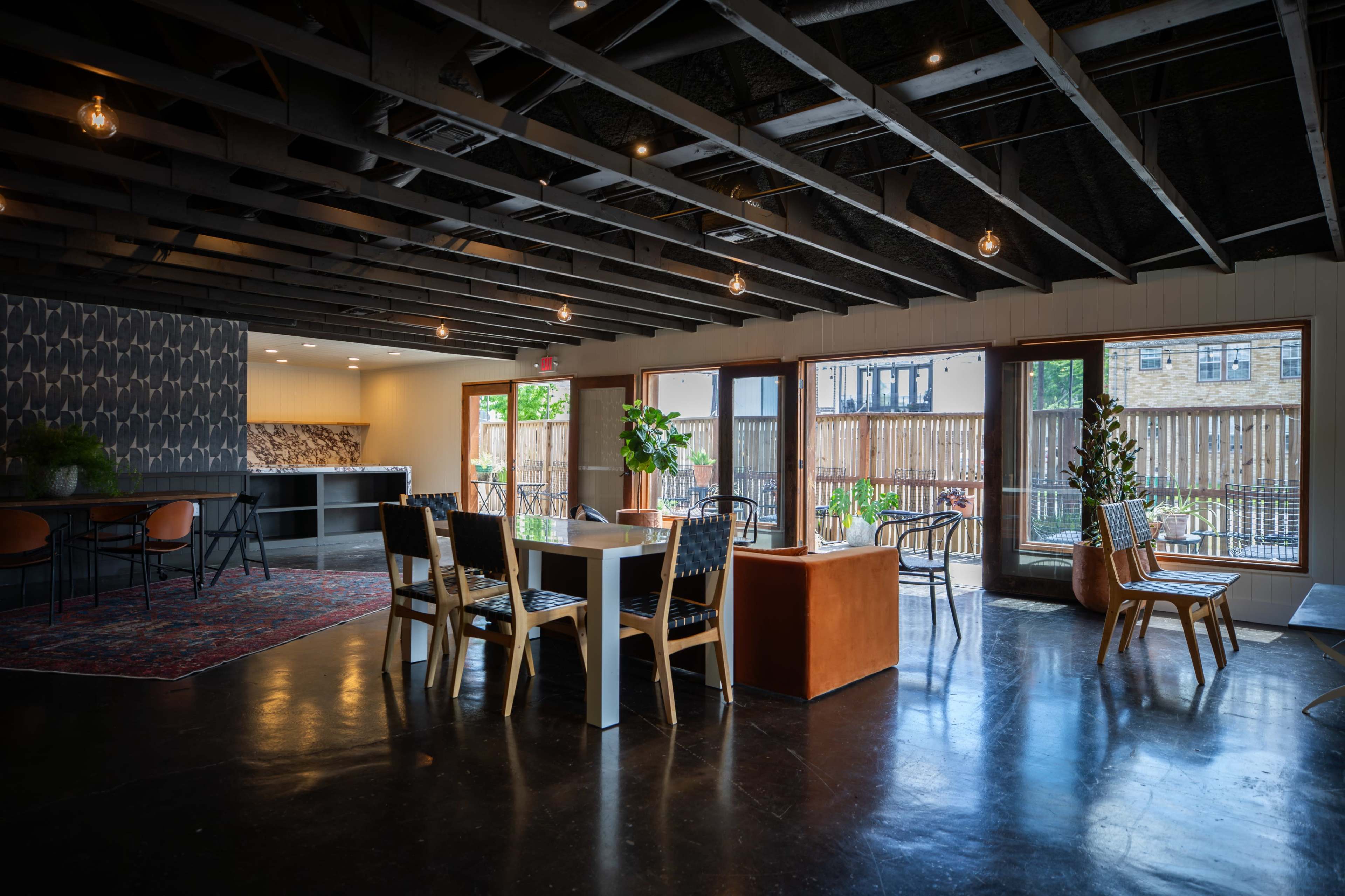 The image shows a modern dining area featuring a large table surrounded by chairs, with large windows providing views of an outdoor space and showcasing a black ceiling with industrial-style lighting.