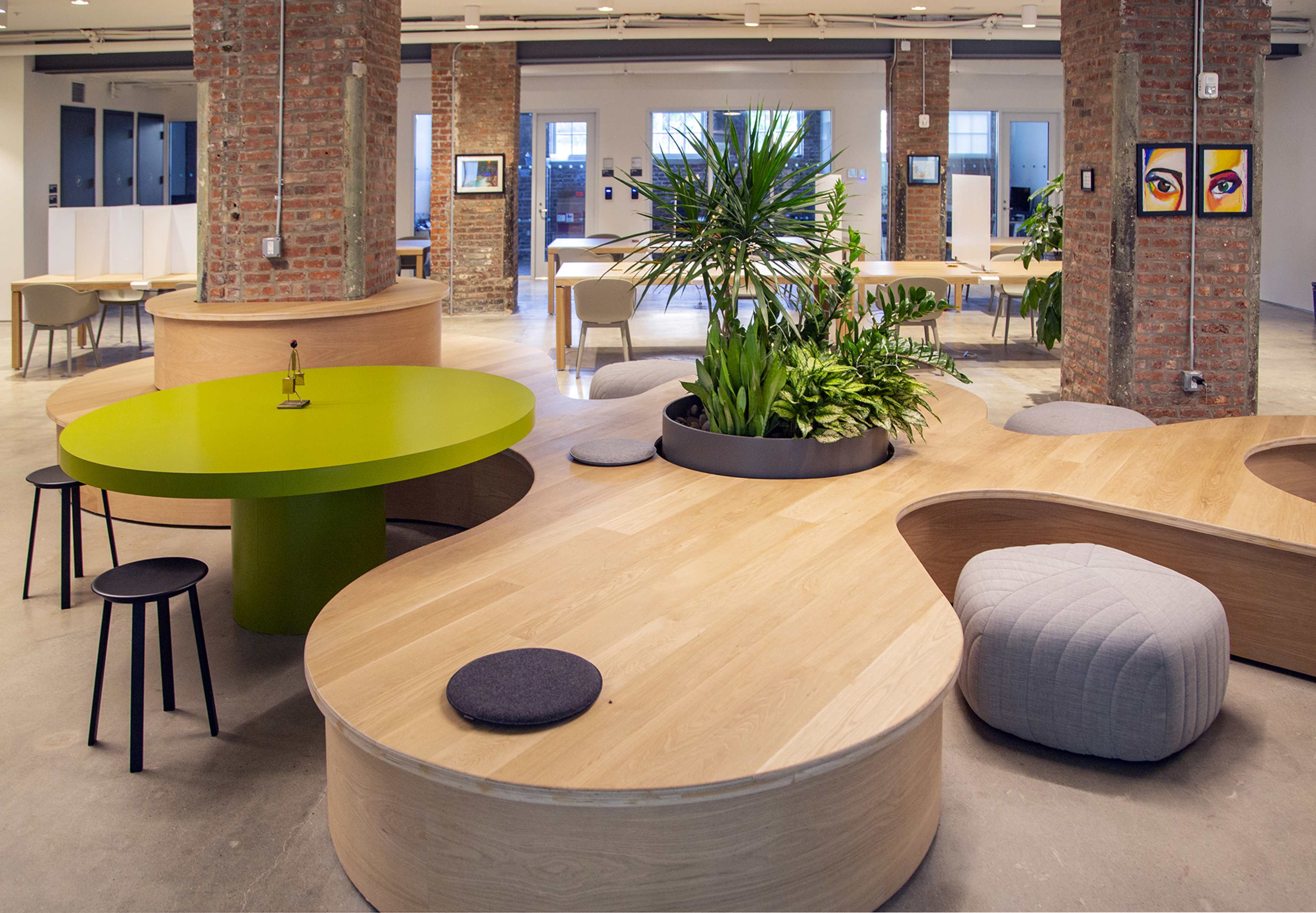 A modern co-working space with wooden tables, circular seating, and a large potted plant at the center.