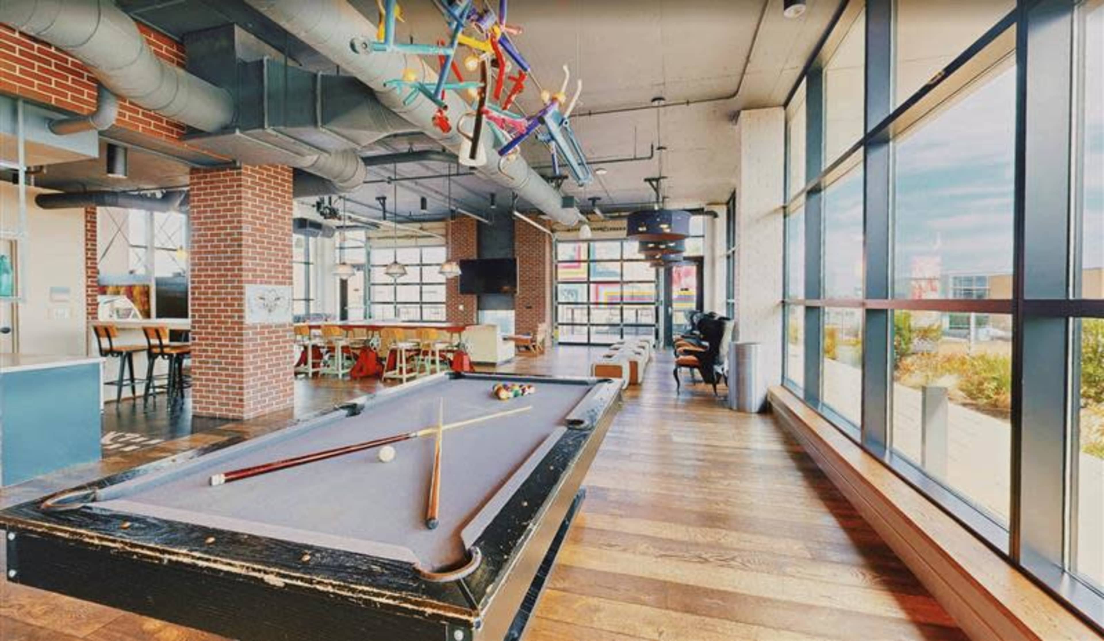 The image shows a modern recreational space featuring a pool table, dining area, and large windows, with colorful decorations hanging from the ceiling.