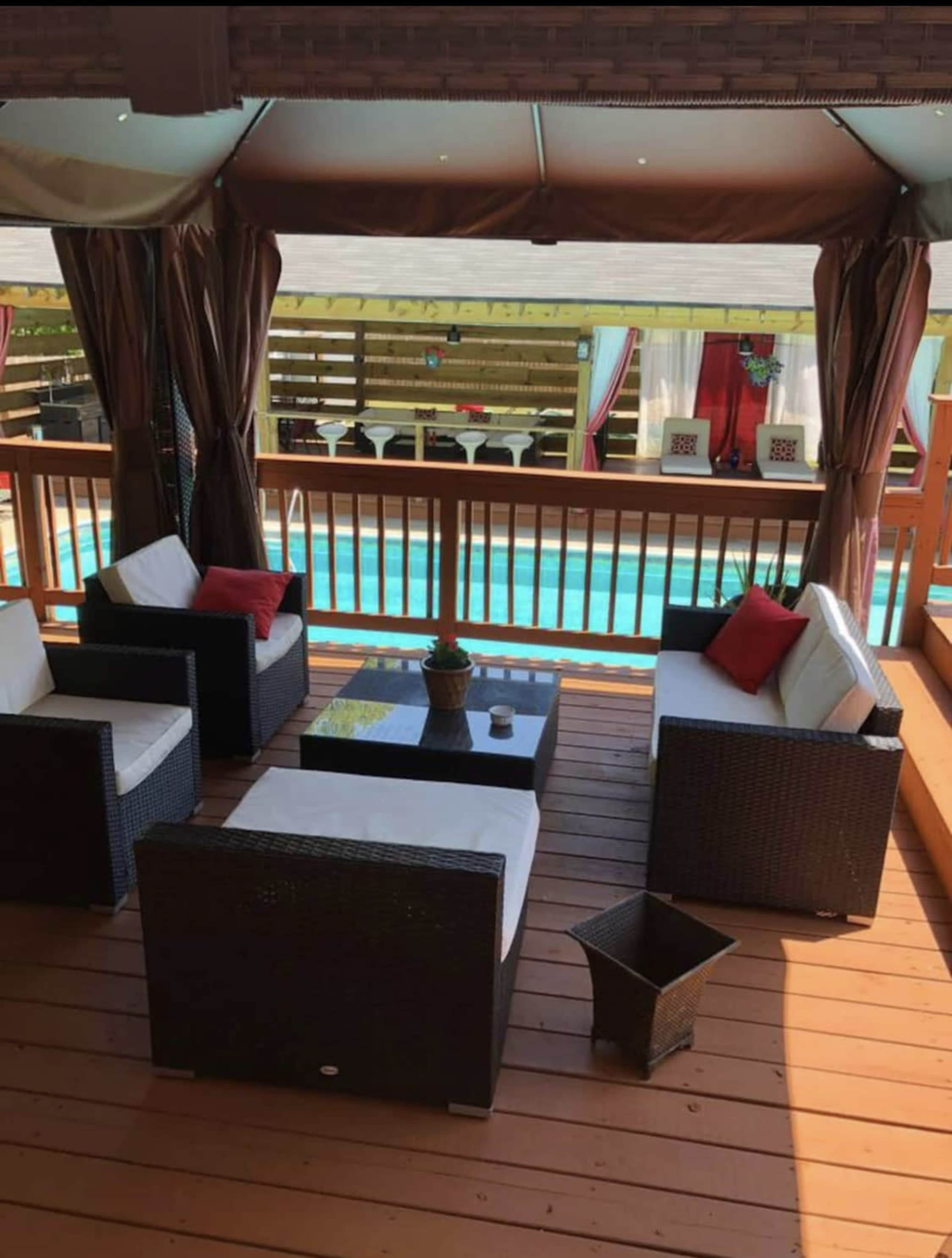 A patio setting features four cushioned chairs around a low table, overlooking a pool area enclosed by a wooden railing.