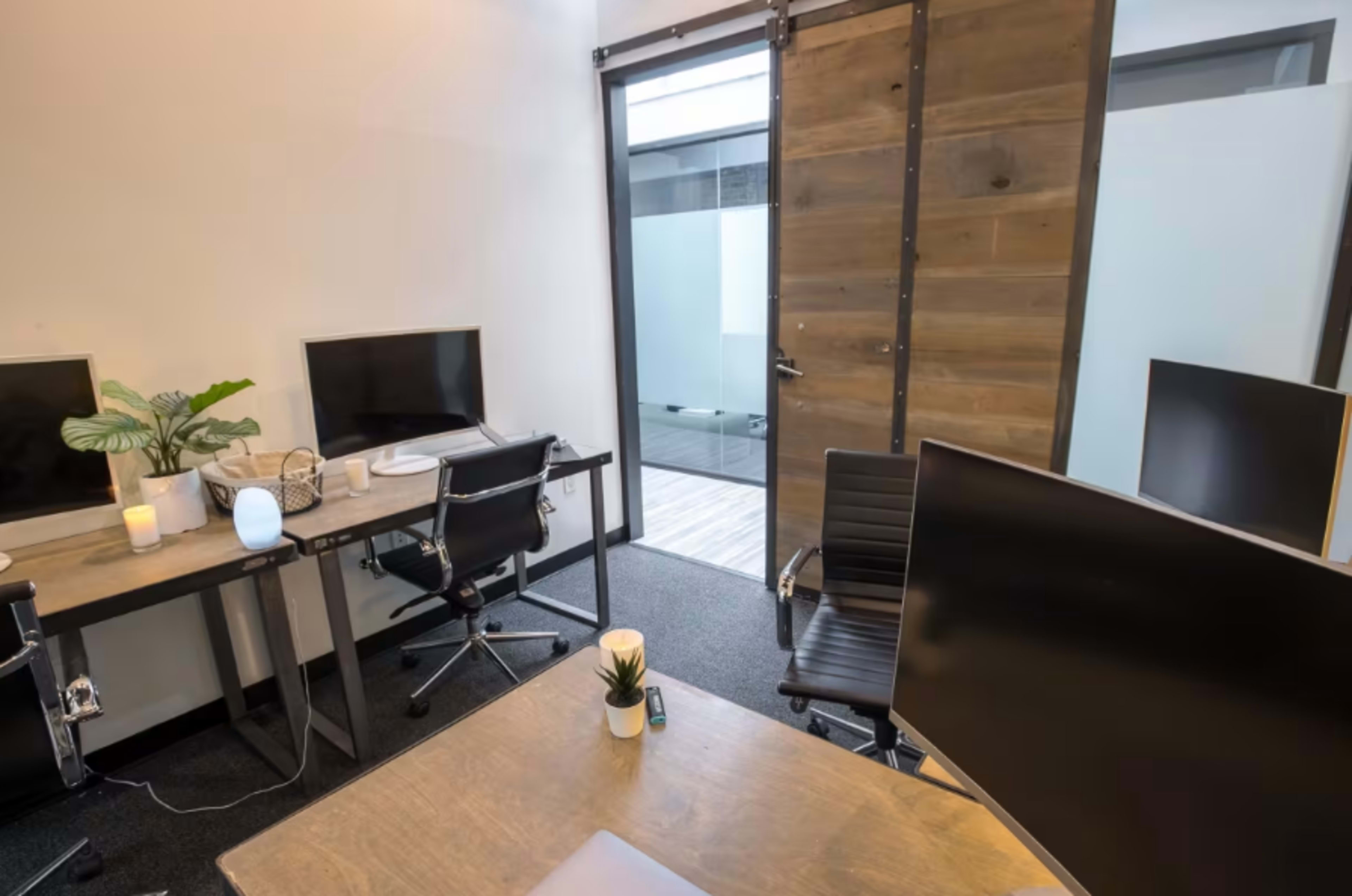 The image shows a modern office space with multiple workstations featuring computer monitors, chairs, and a door leading to a glass-enclosed area.