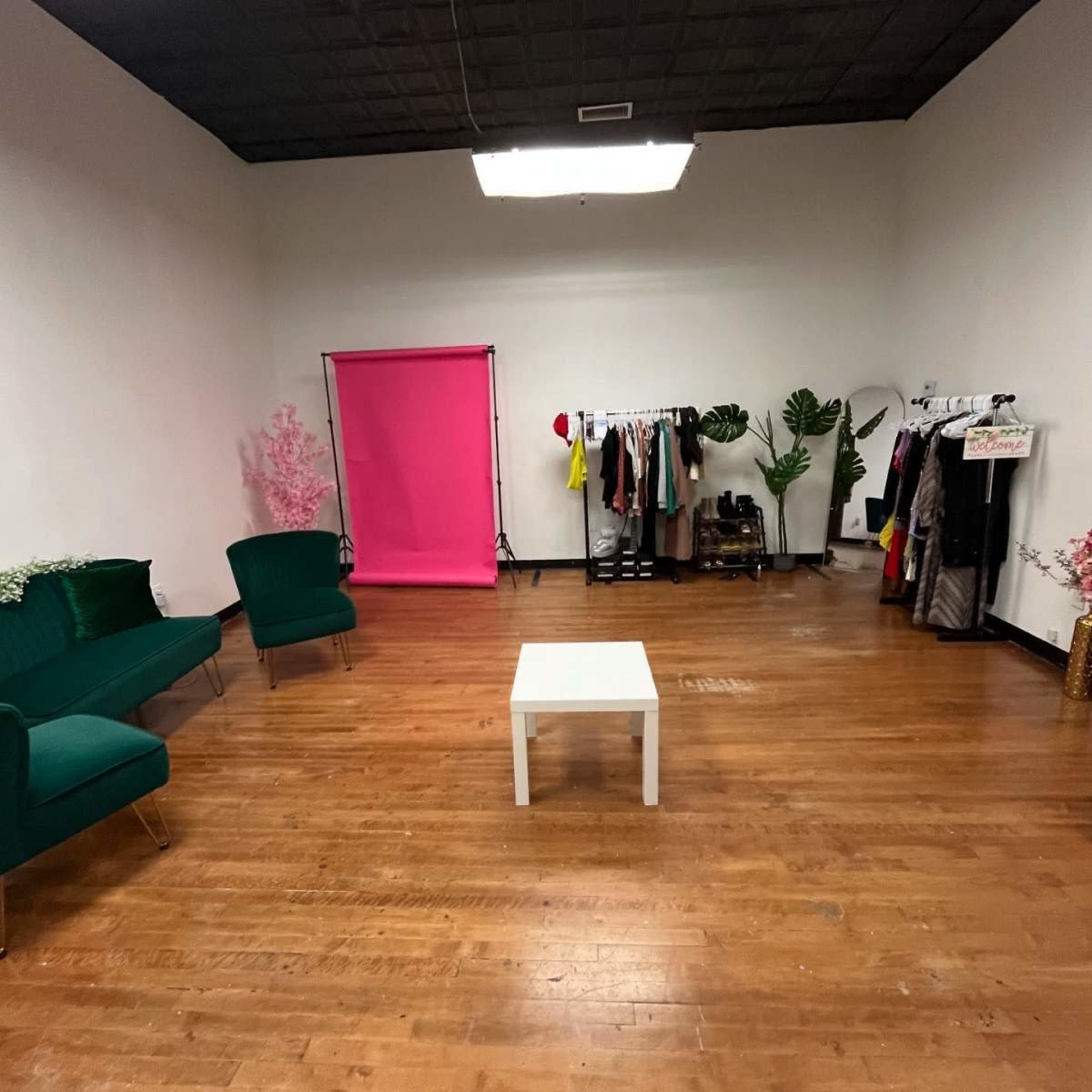The image shows a spacious room with hardwood flooring, featuring a pink backdrop, clothing rack, a small white table, and two green chairs.
