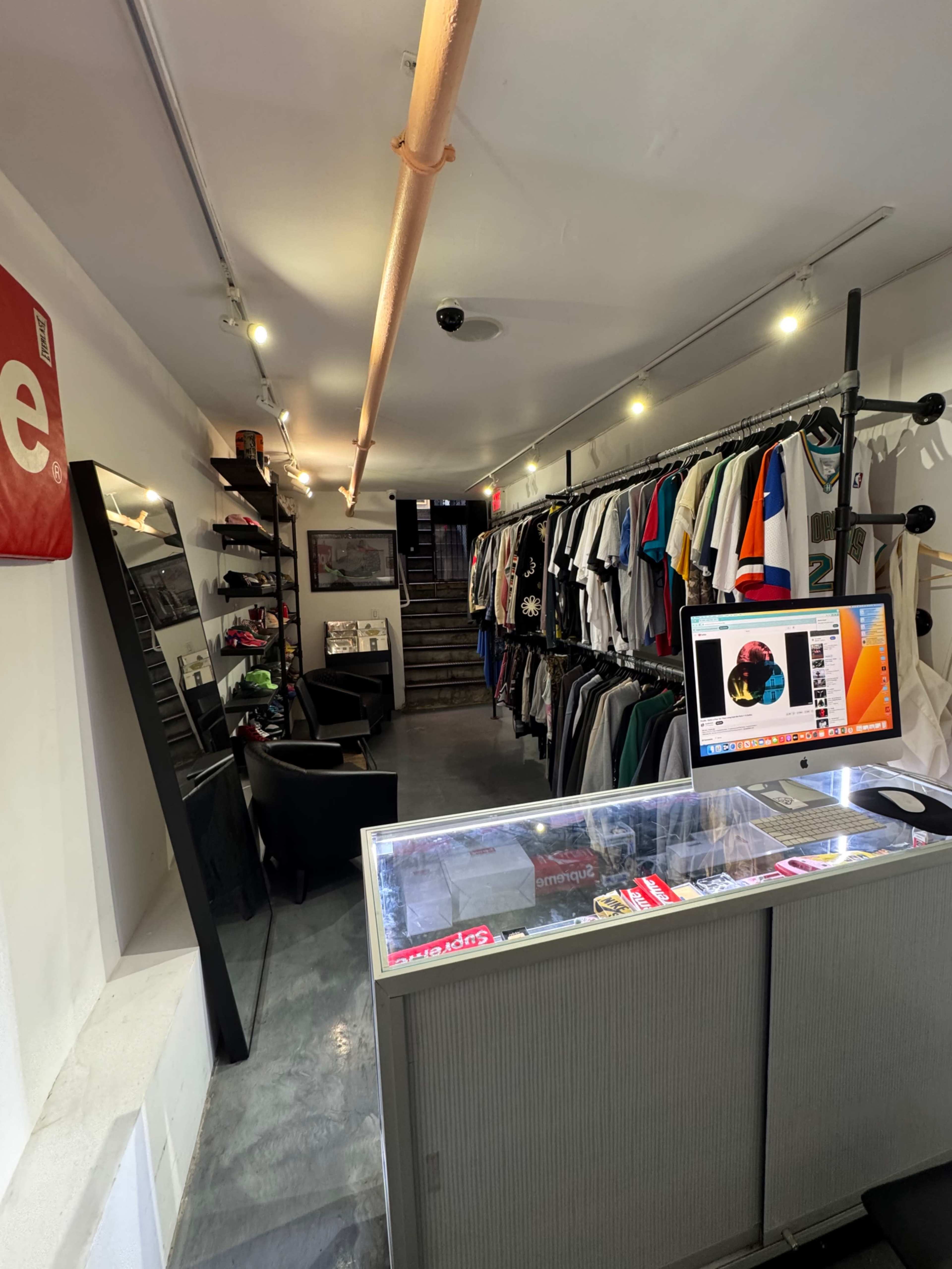 The image shows an interior of a retail store with clothing racks on one side and a checkout counter with a computer on the other, leading to a staircase in the background.