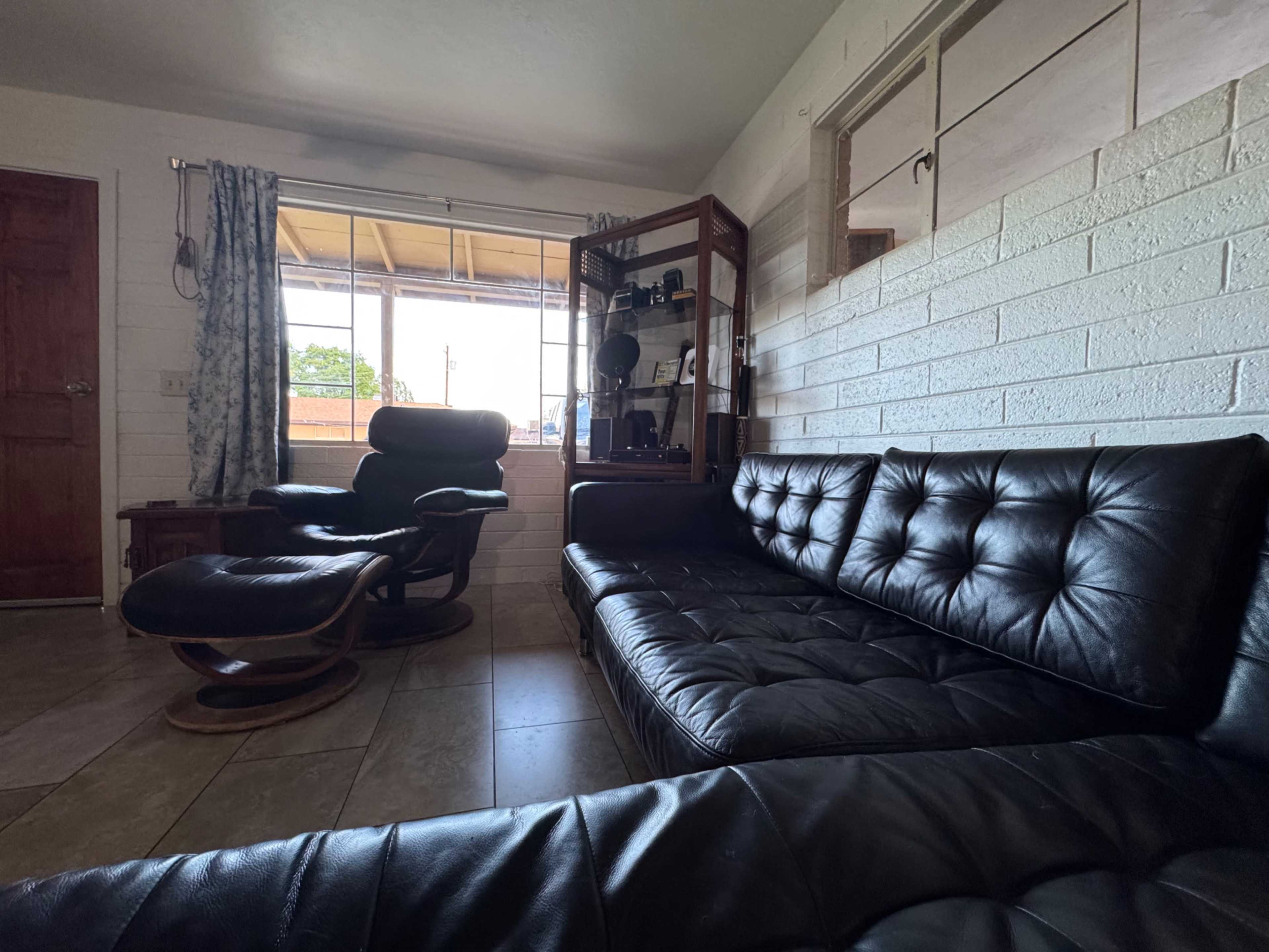The image shows a living room with a black leather sectional sofa, a wood accent chair, and a display cabinet, with natural light coming in from a window.