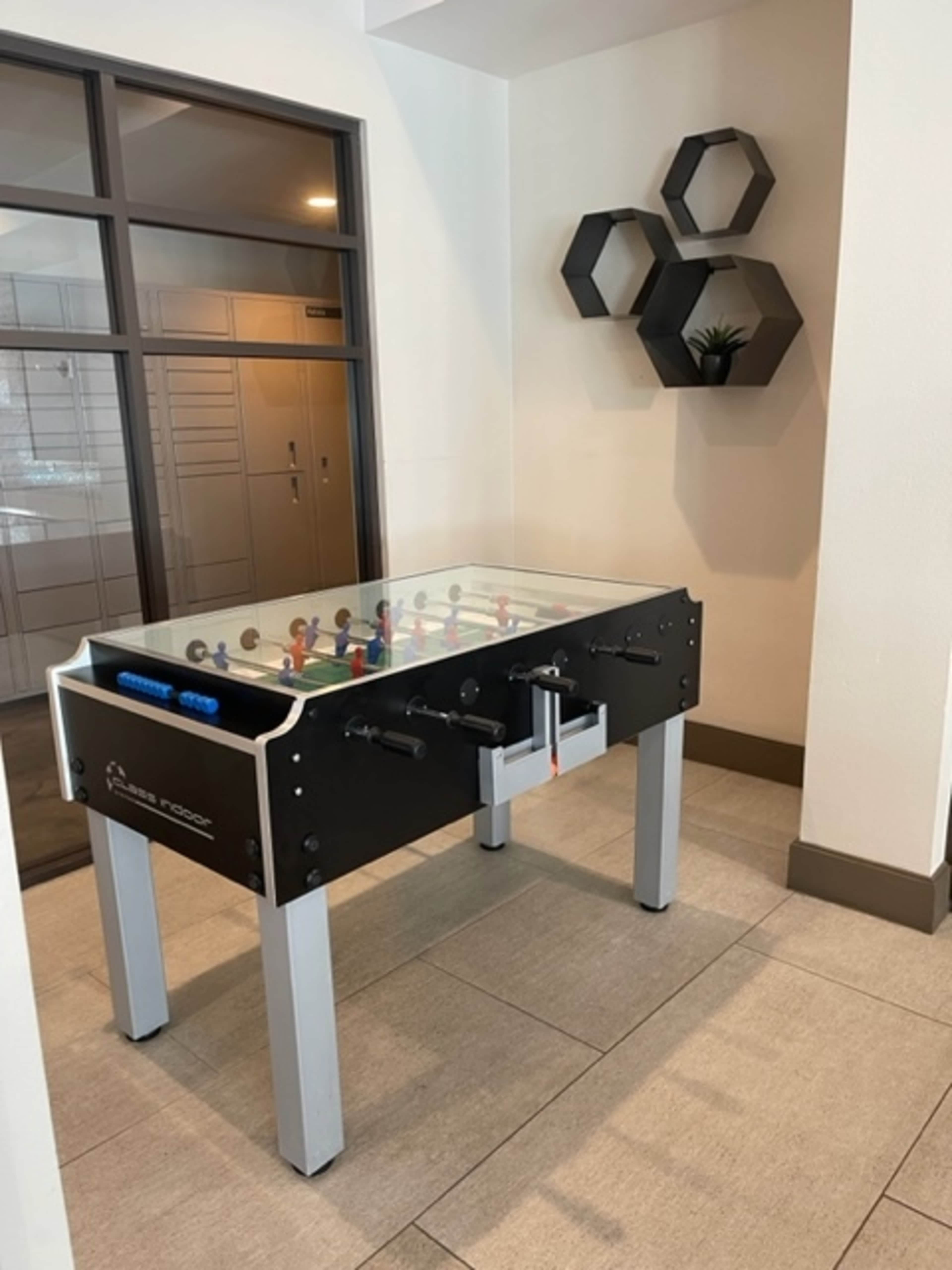 A foosball table is positioned in a corner of a lobby area, adjacent to a wall with decorative hexagonal shelves.