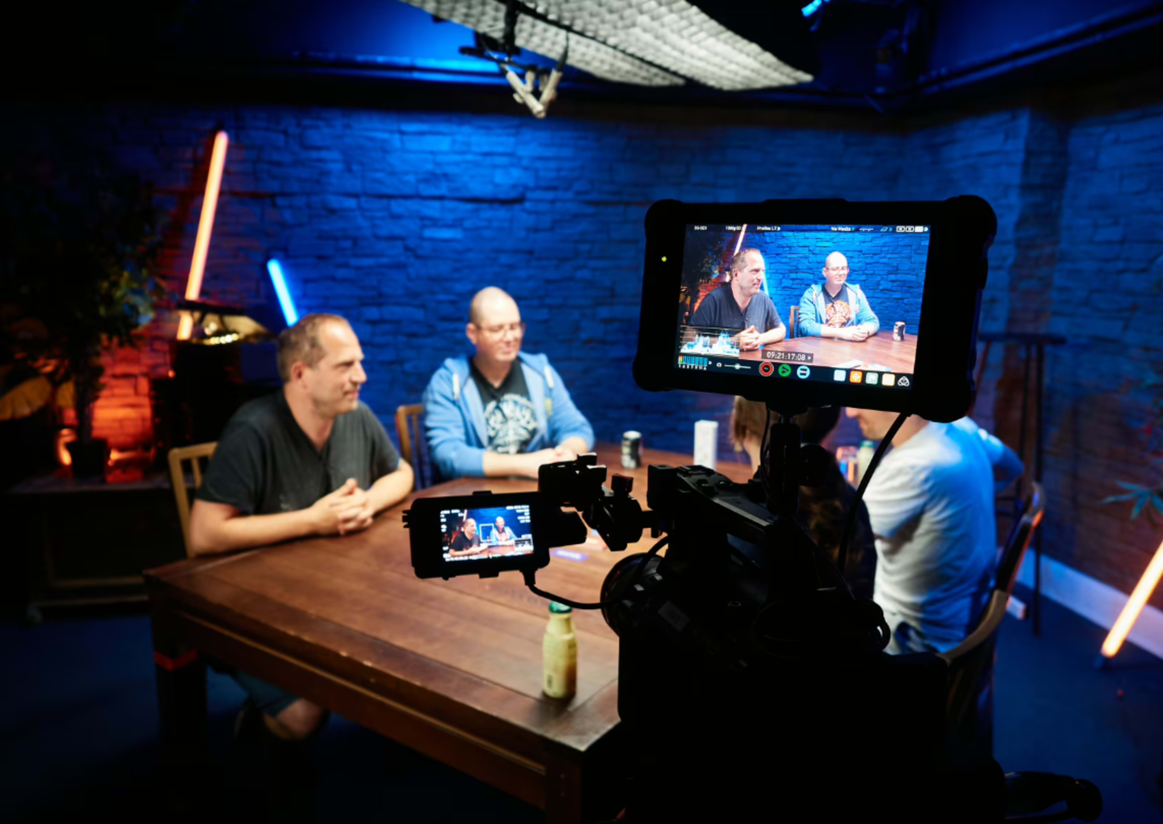 A group of four people sits around a table in a dimly lit room with colorful lights, while cameras are positioned to capture the conversation.