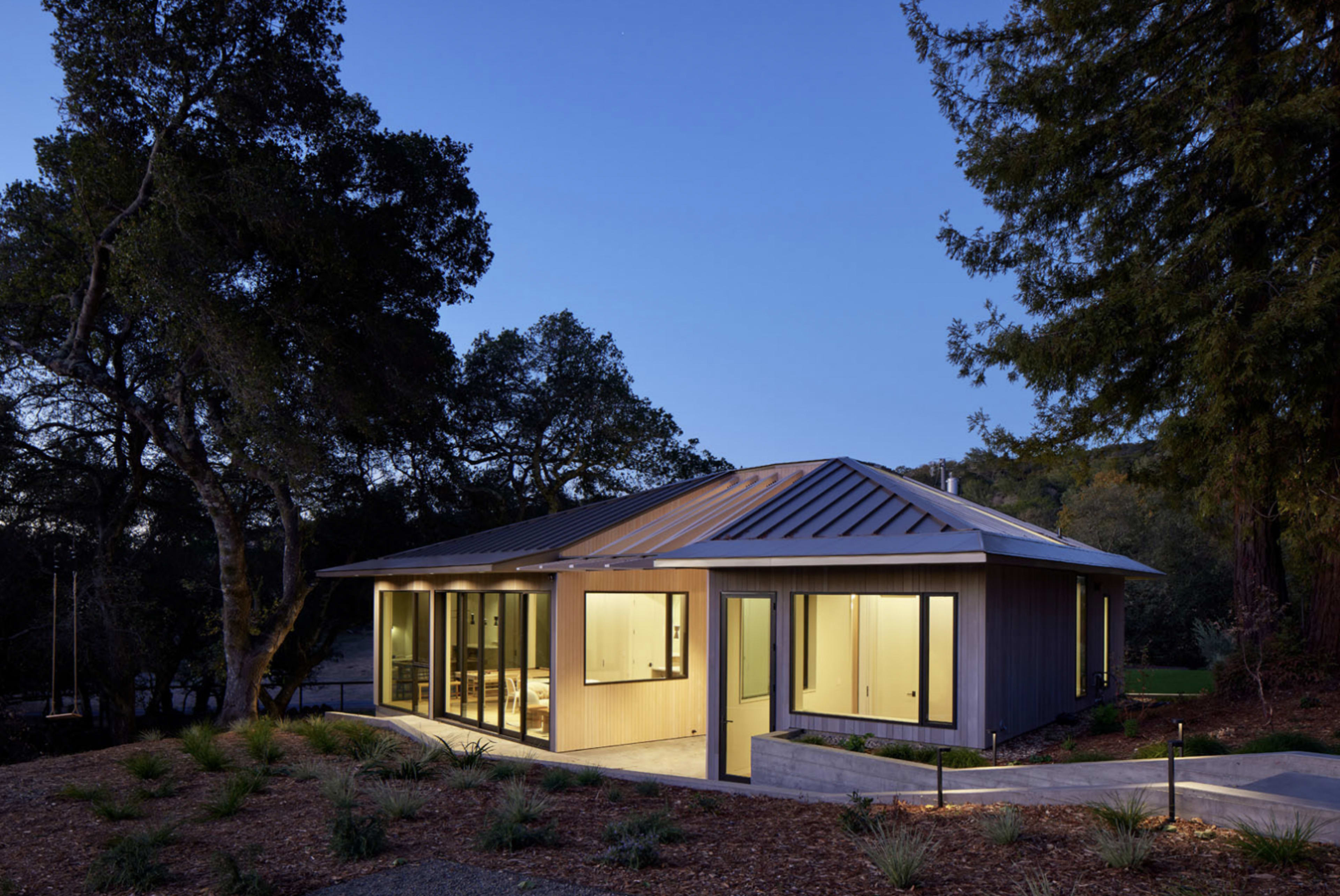 A modern single-story house with a metal roof and large glass windows is situated among trees in the evening light.