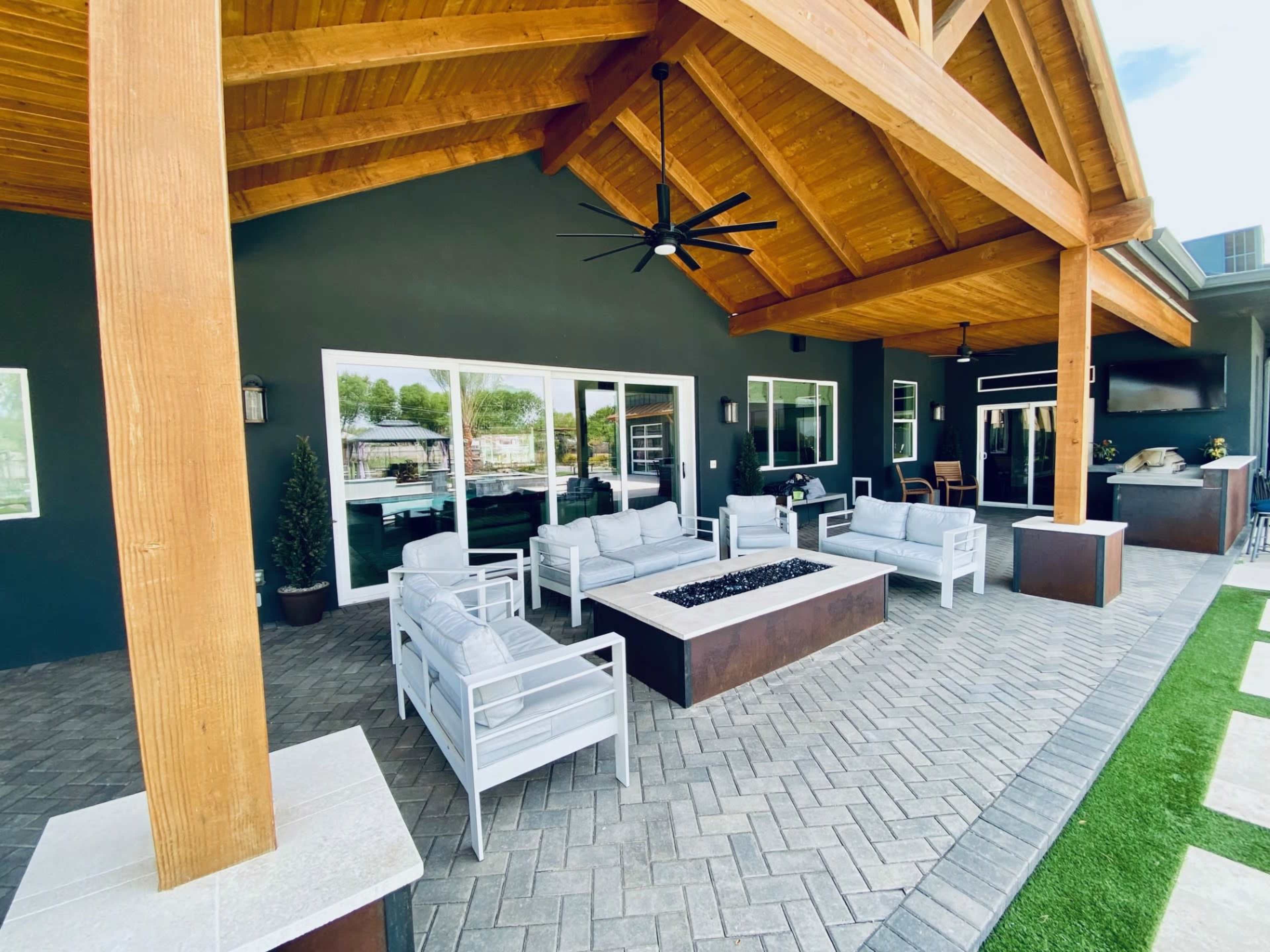 The image shows a patio area with a wooden ceiling, modern outdoor furniture arranged around a fire pit, and large windows overlooking a landscaped space.