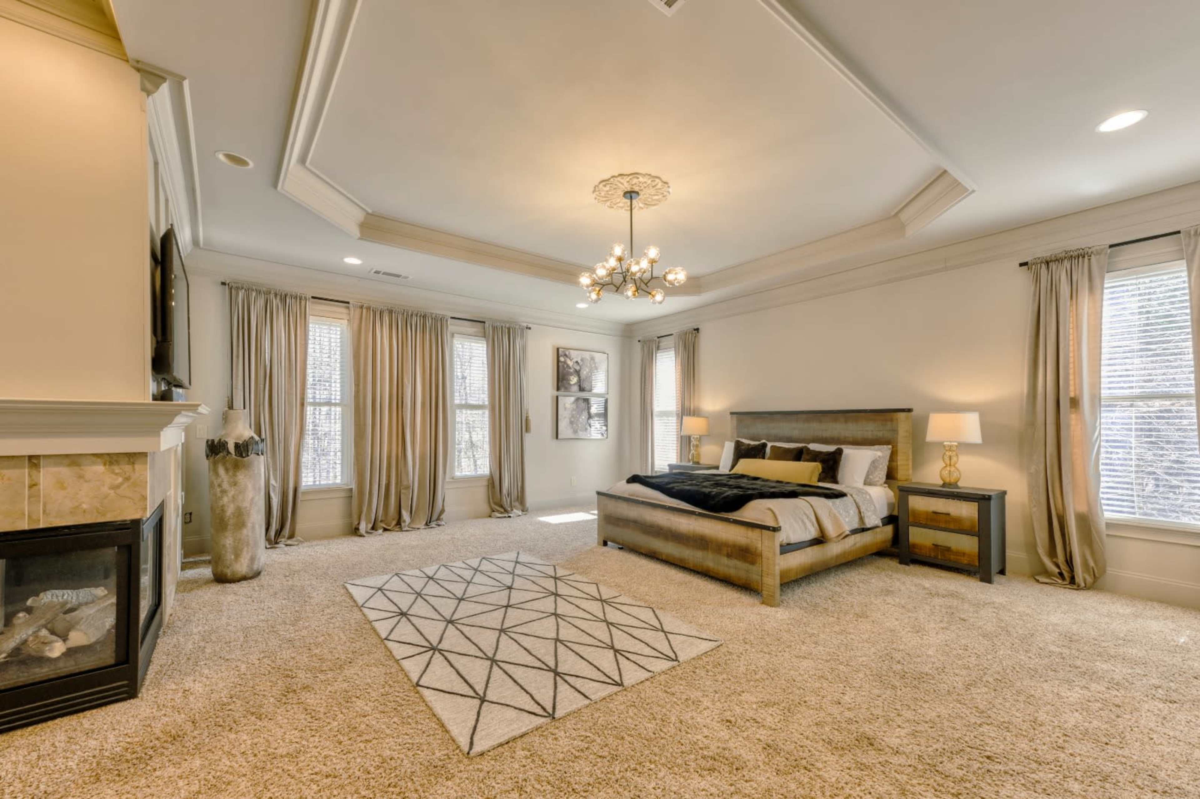 A spacious bedroom featuring a bed with dark bedding, a nightstand with a lamp, a fireplace, and large windows covered with beige curtains.